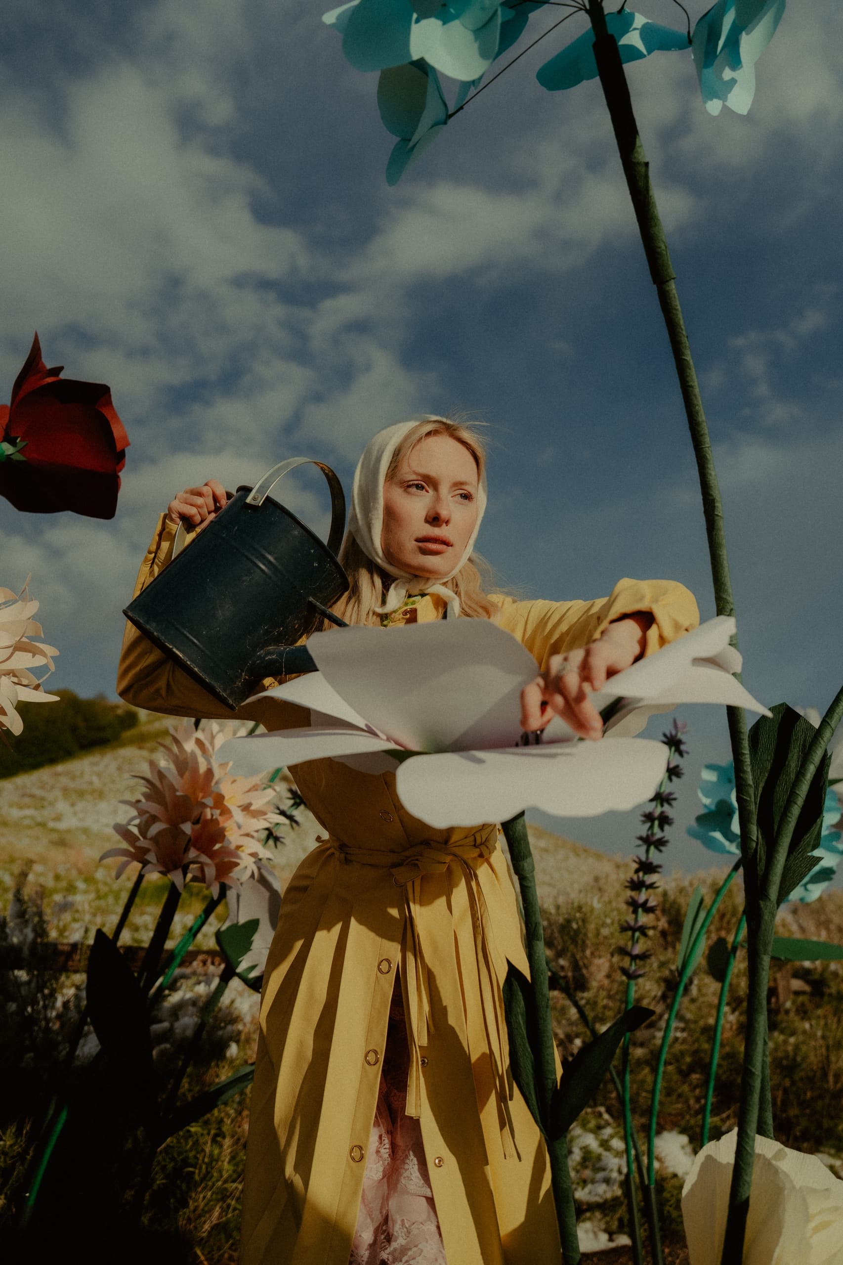 Woman in yellow coat watering oversized flowers in a surreal garden scene.