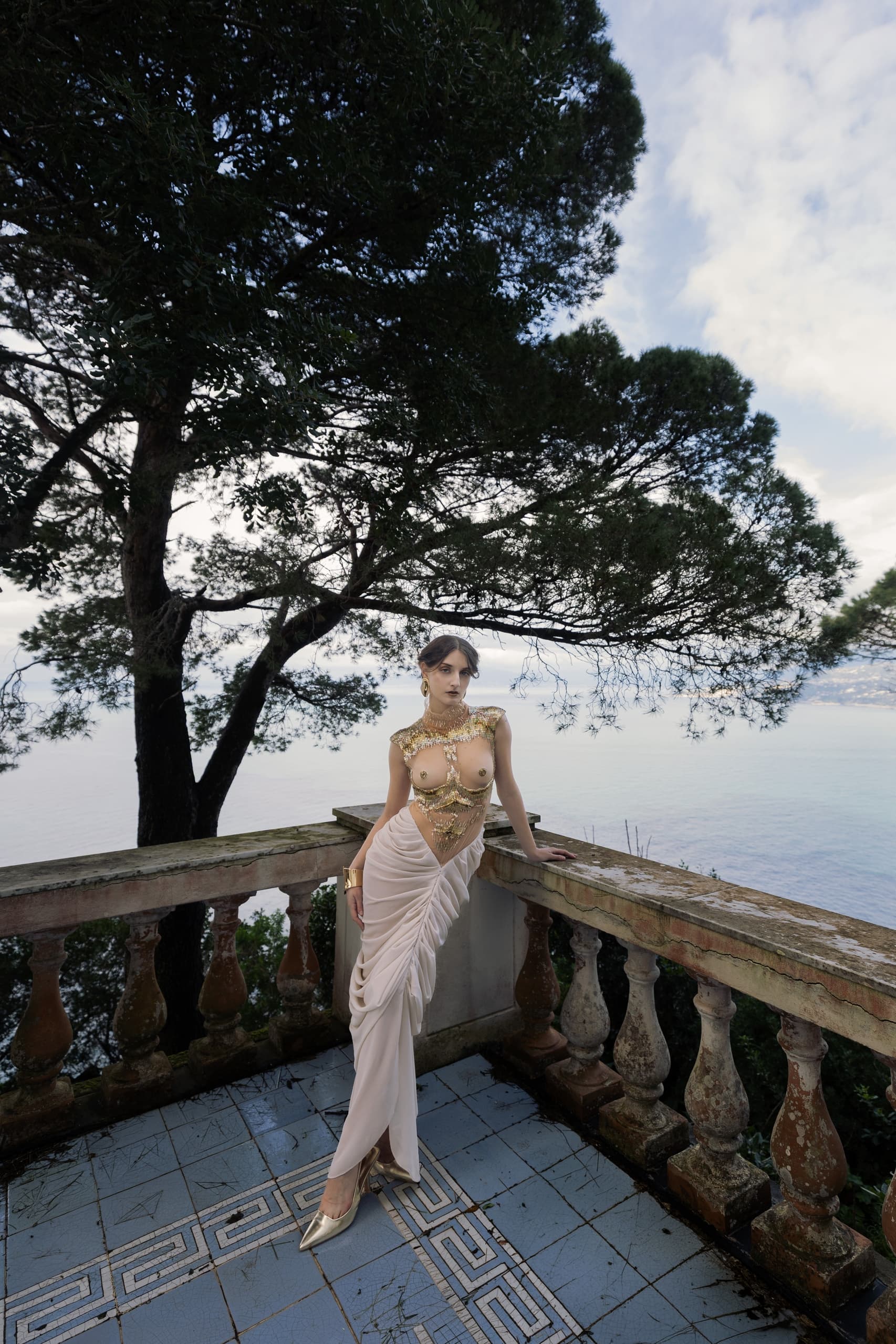 Elegant woman in a stylish gown on a seaside terrace surrounded by trees.