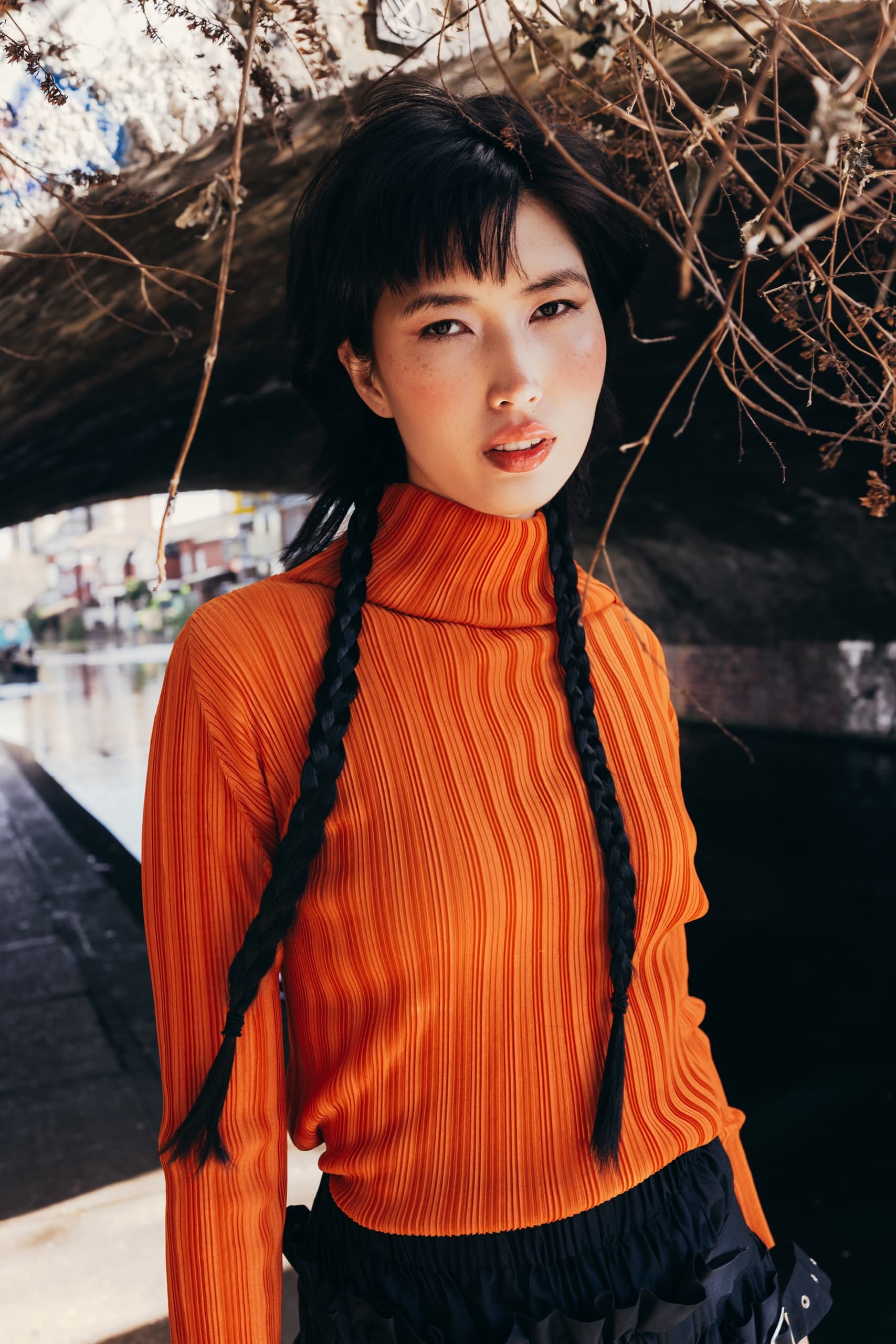 Person in orange sweater with braids stands under a bridge with dried branches overhead.