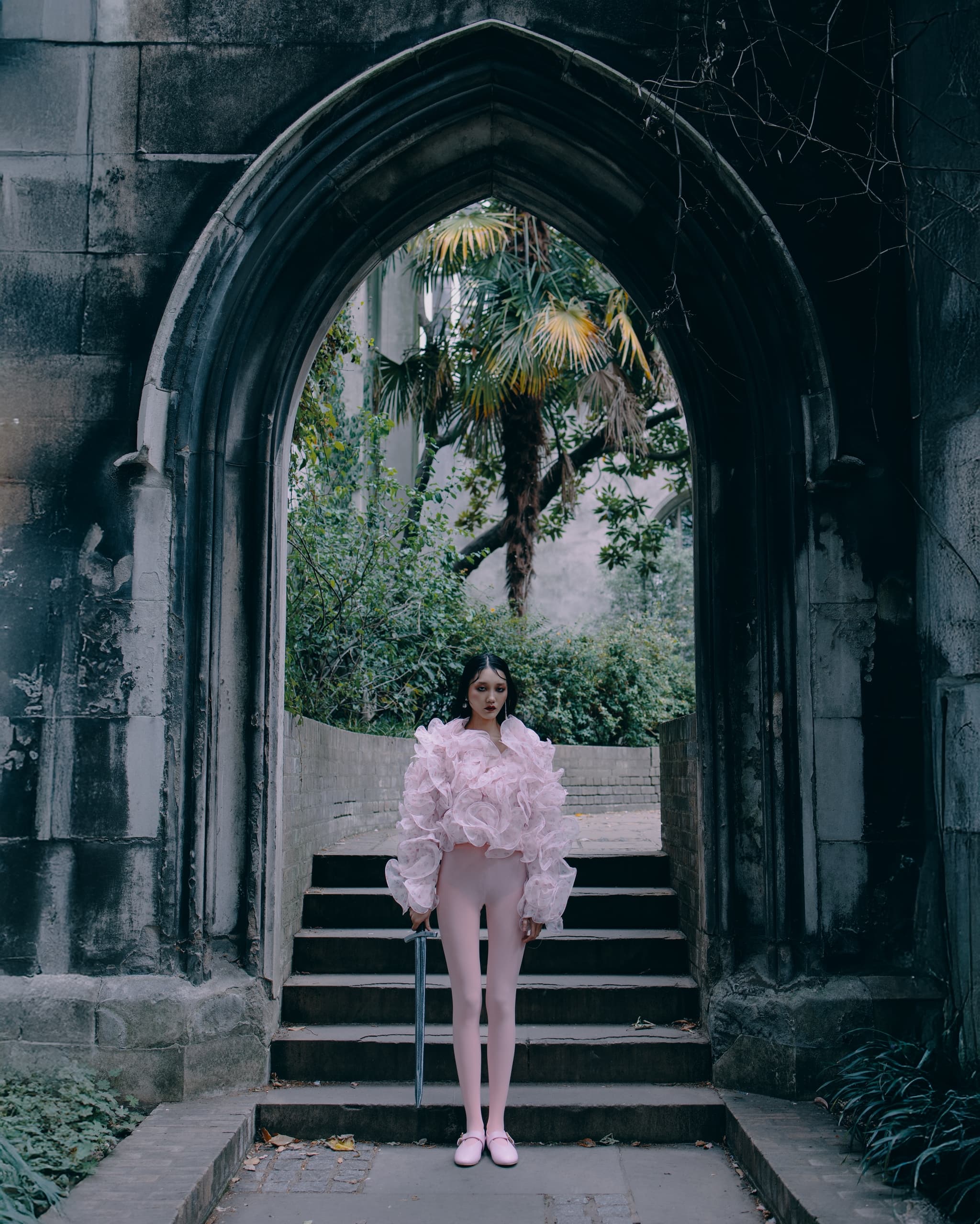 Person in elaborate pink outfit standing in gothic archway with lush greenery.