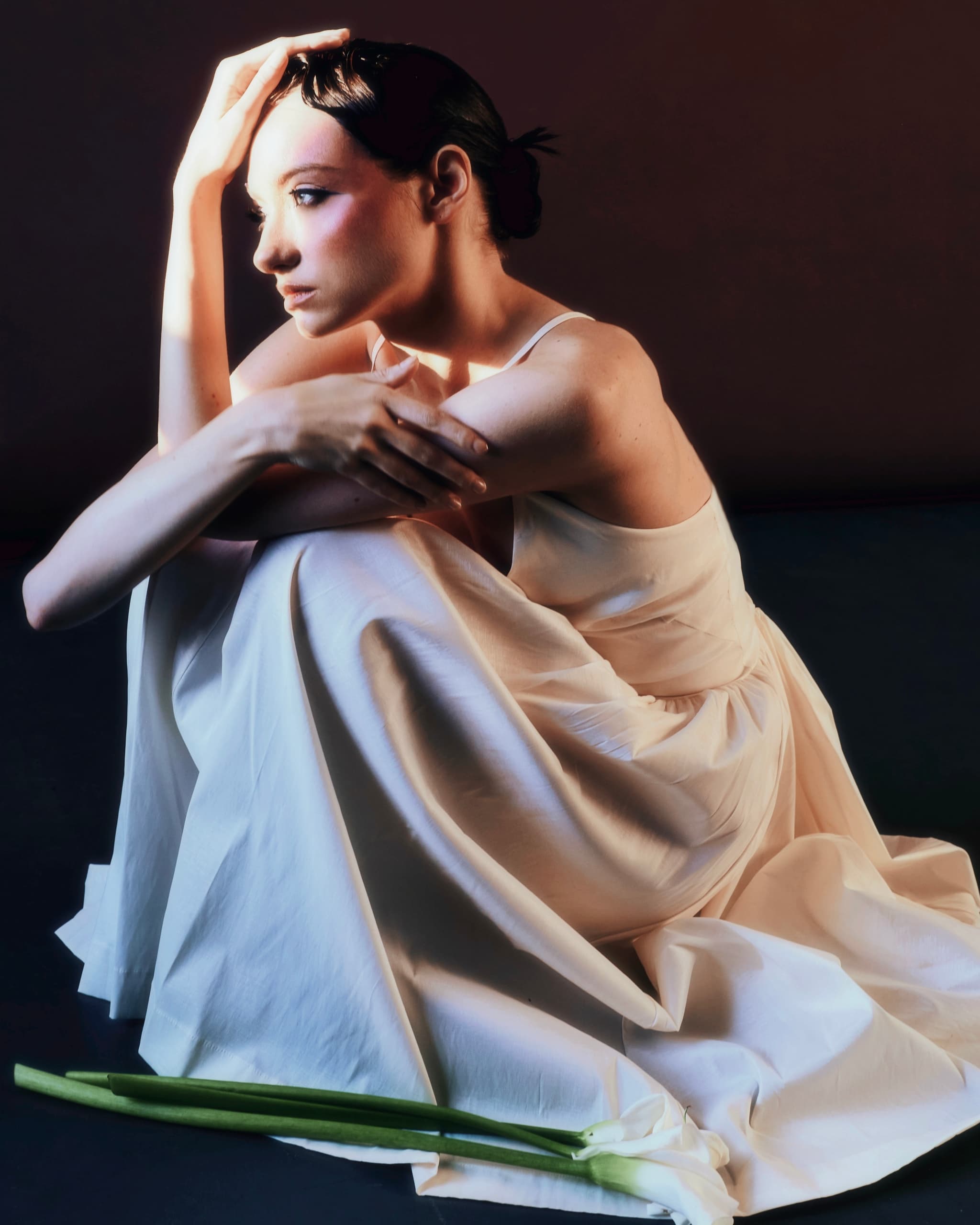 Woman in elegant white dress sitting thoughtfully, with a lily on a dark background.