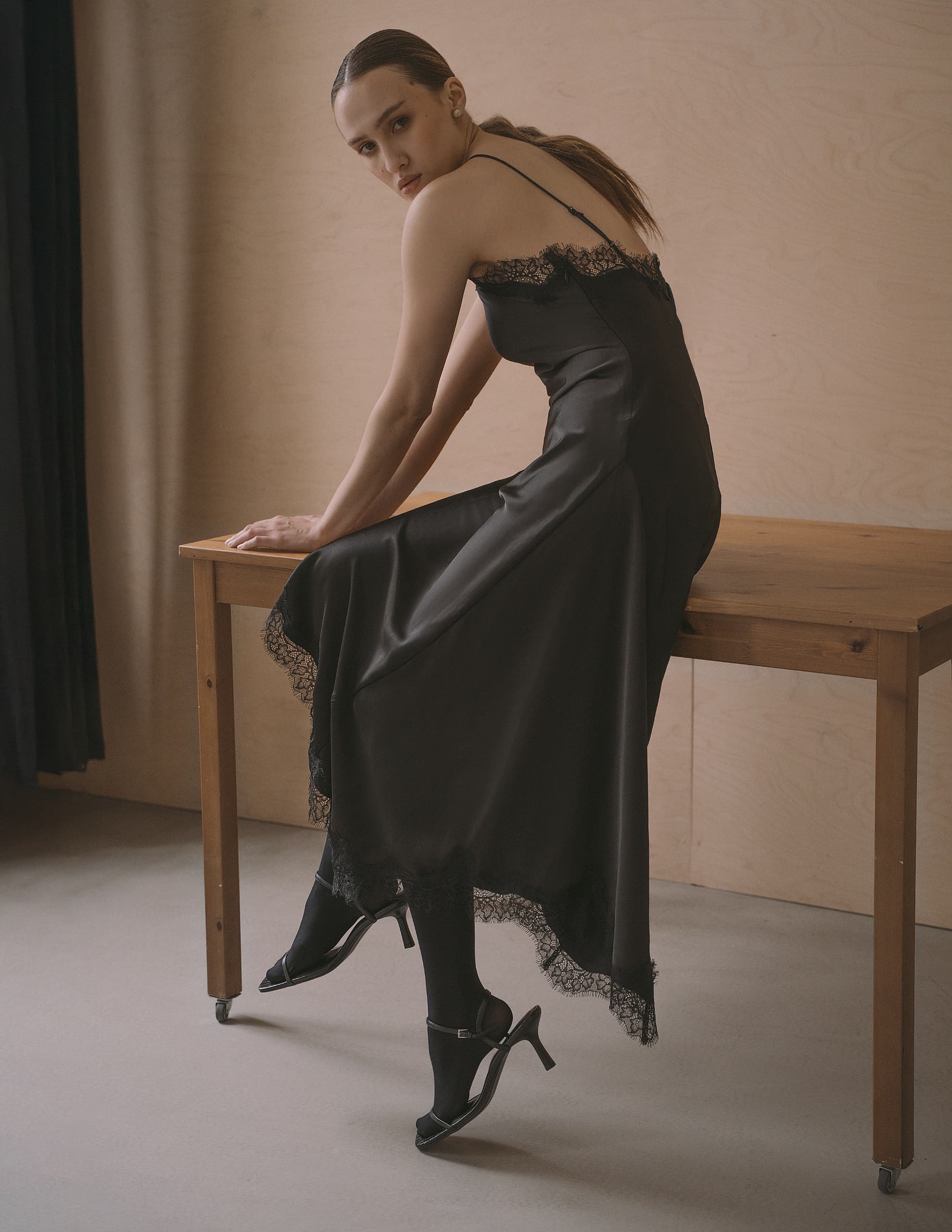 Elegant woman in a black dress sitting on a table against a minimalist background.