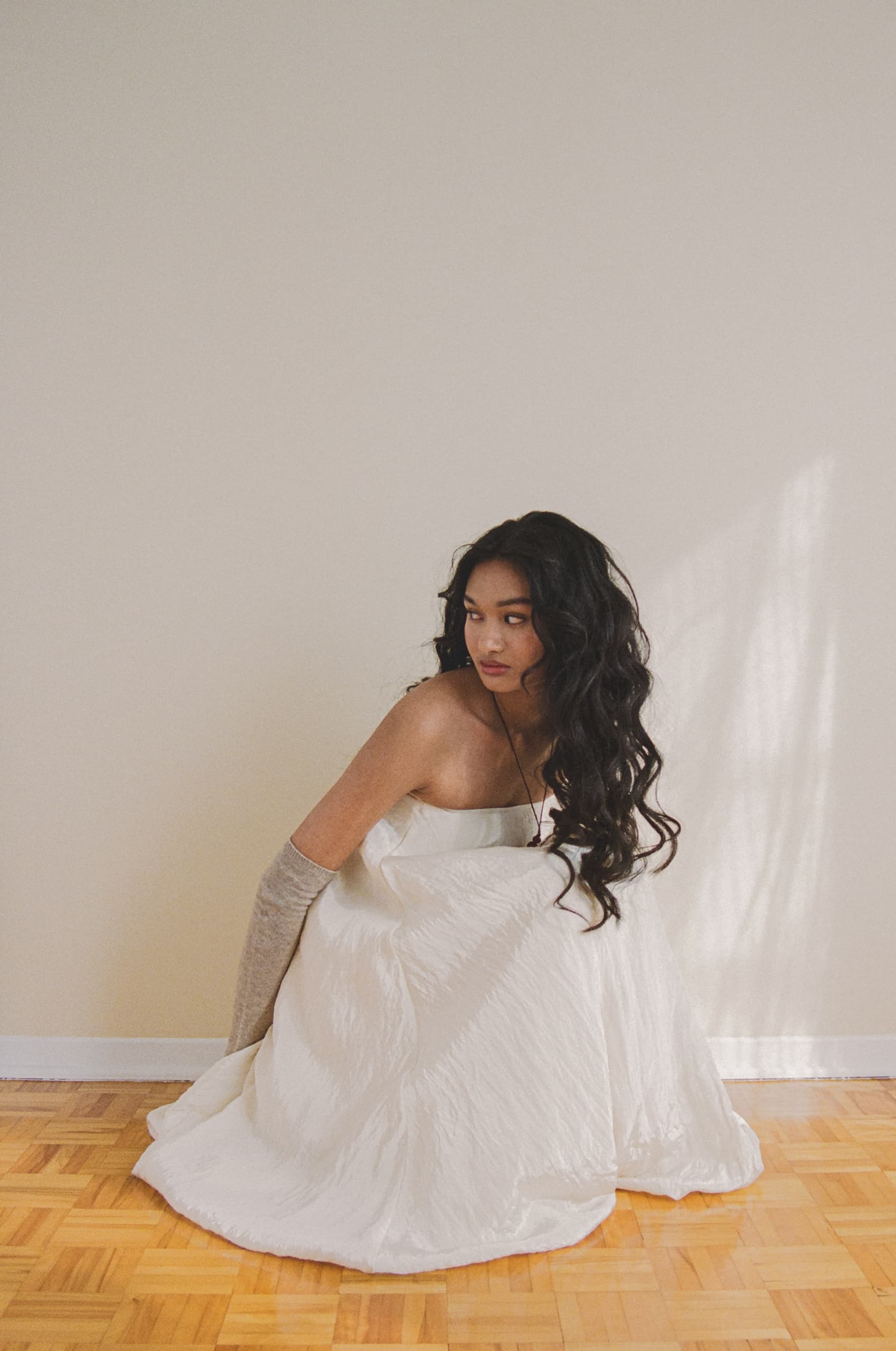 Woman in elegant white dress sitting on parquet floor, soft lighting.