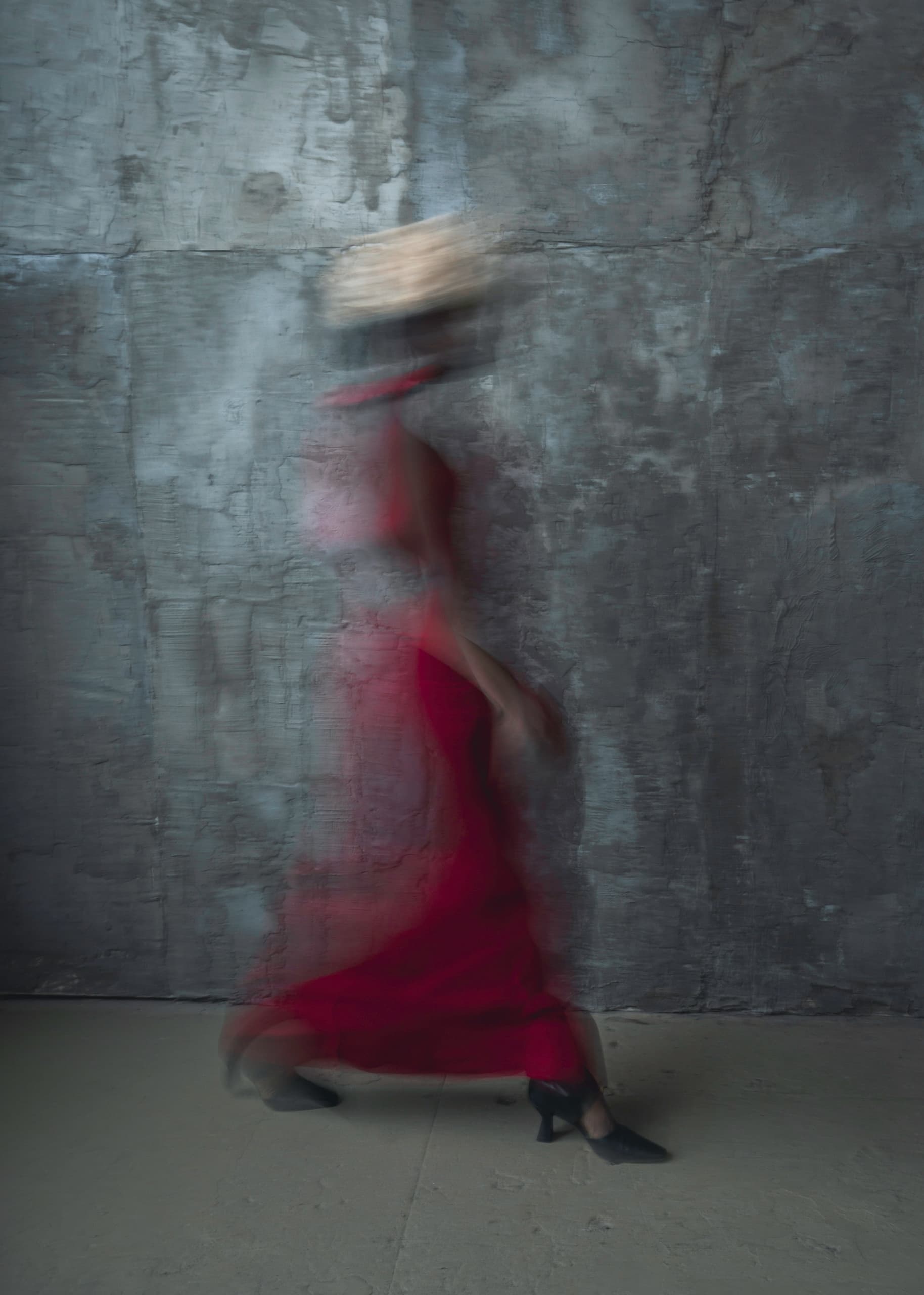 Blurred silhouette in motion, wearing a red dress against a textured gray wall.