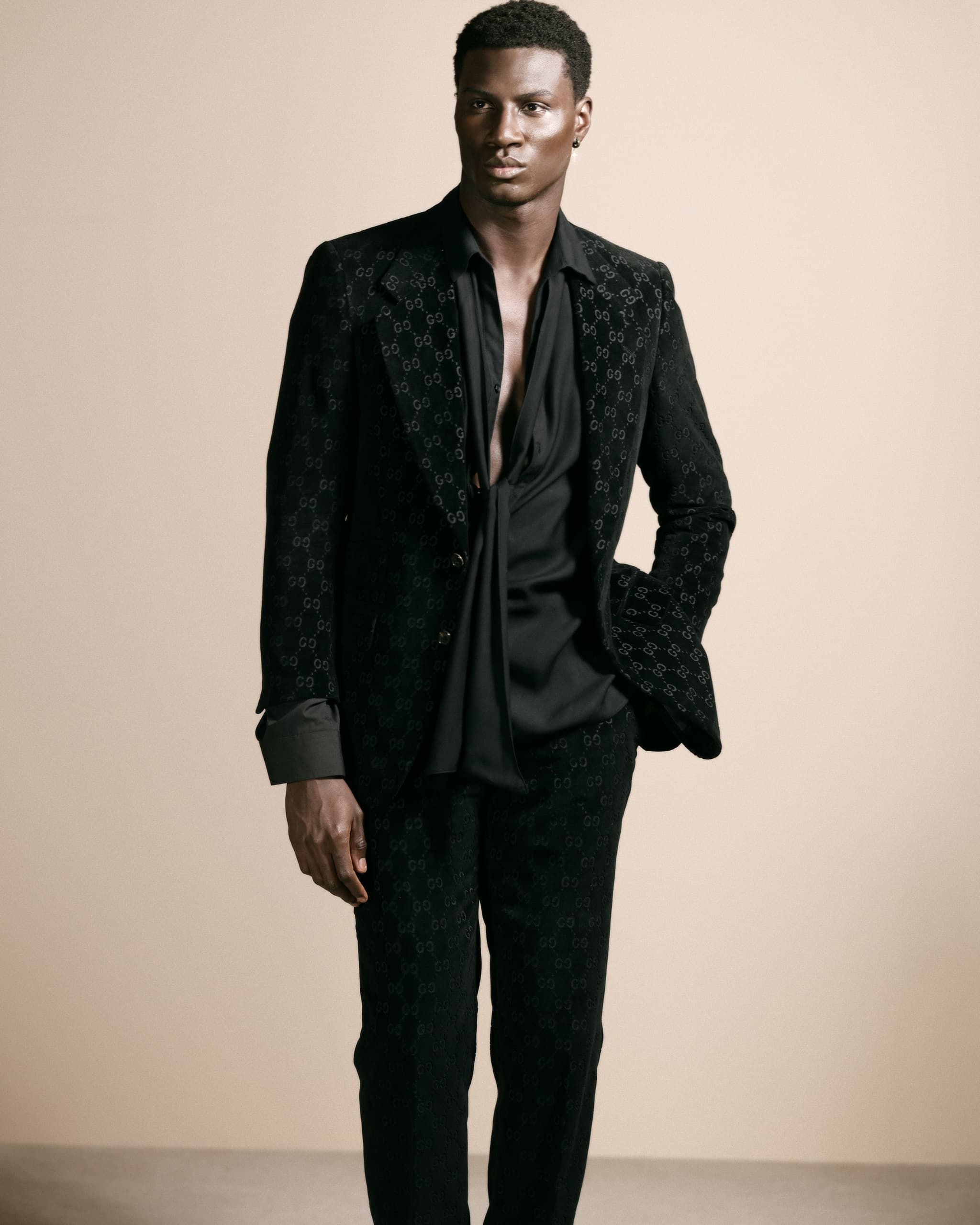 Man in elegant black suit with patterned design, posing confidently on plain background.