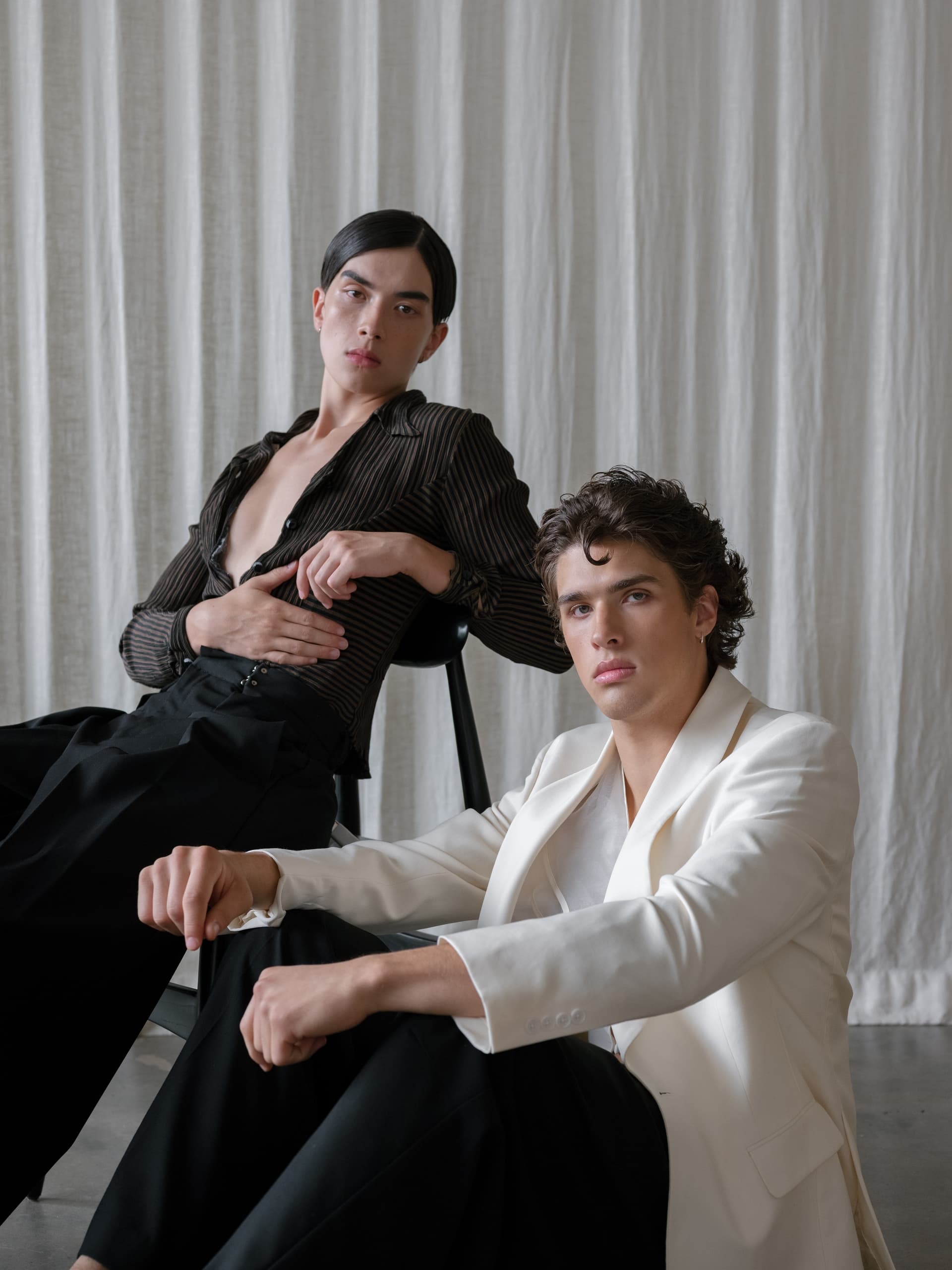 Two stylish models posing in chic, monochrome outfits with a soft fabric backdrop.