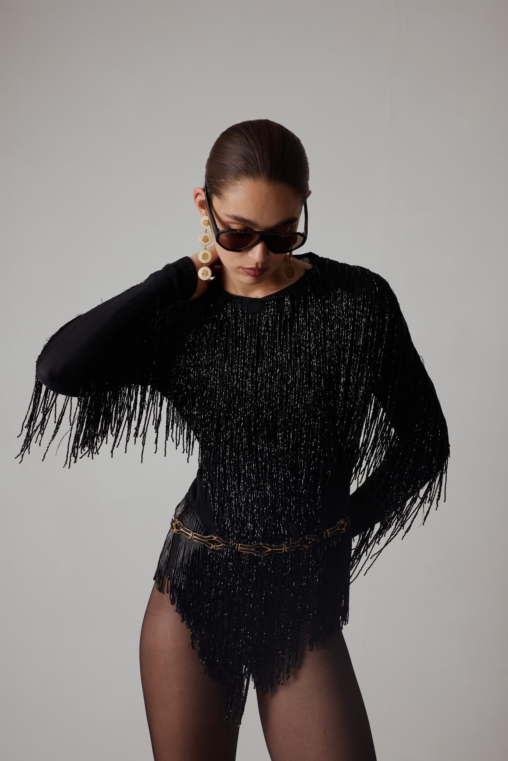 Stylish woman in black fringe outfit, sunglasses, bold earrings, posing confidently.