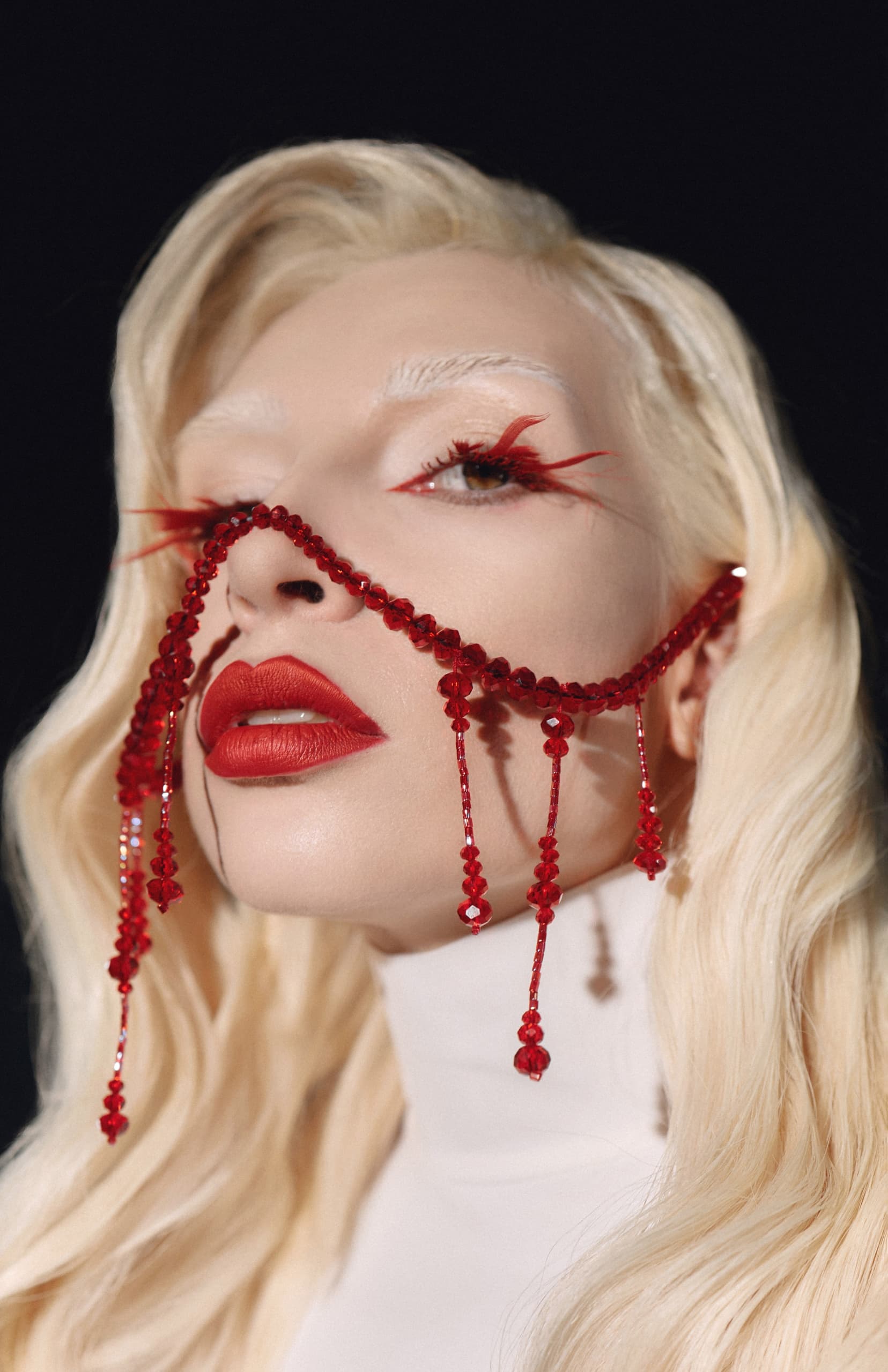 Blonde model with red beaded face adornment and striking makeup in artistic portrait.