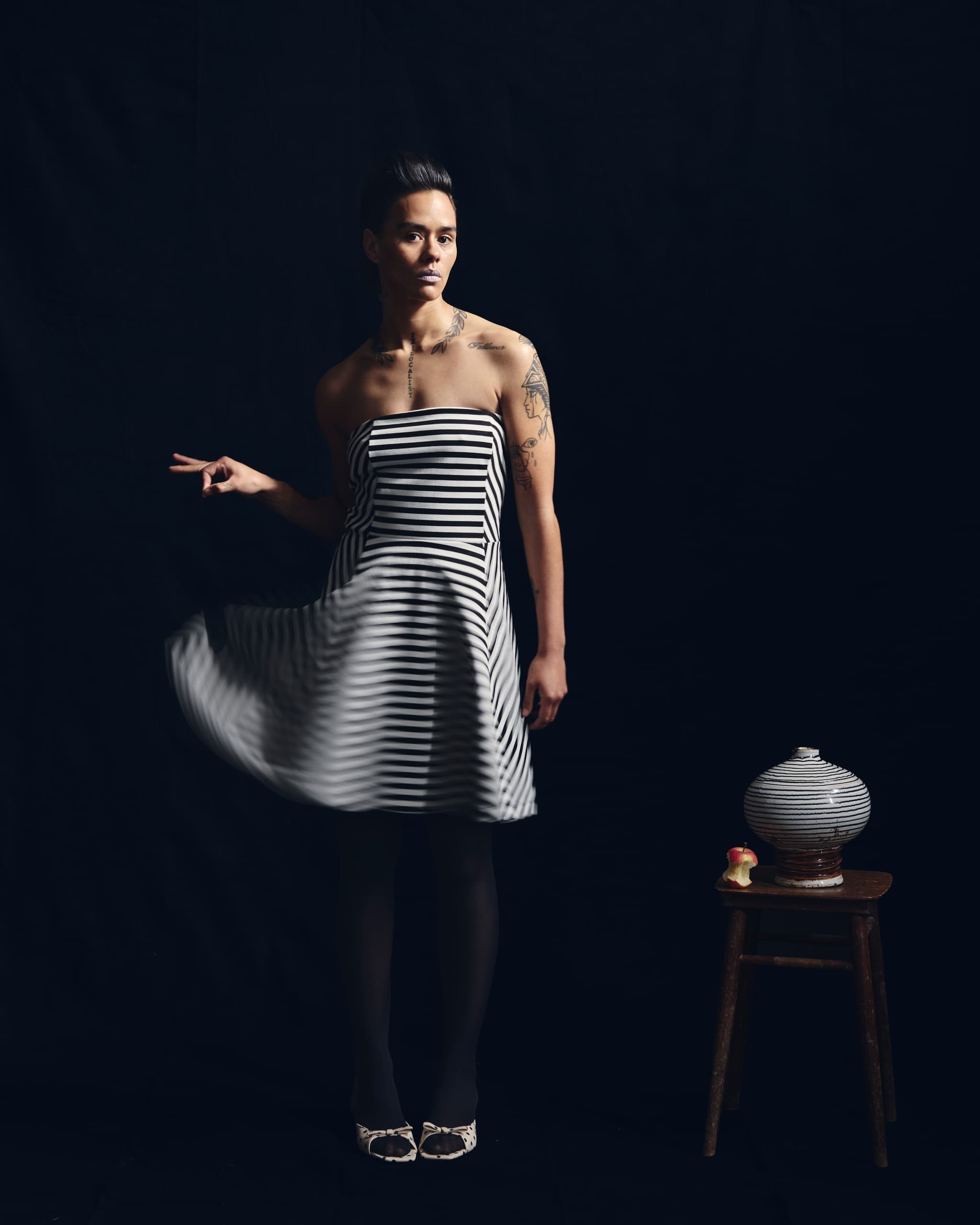 Person in striped dress poses beside stool with vase and apple.