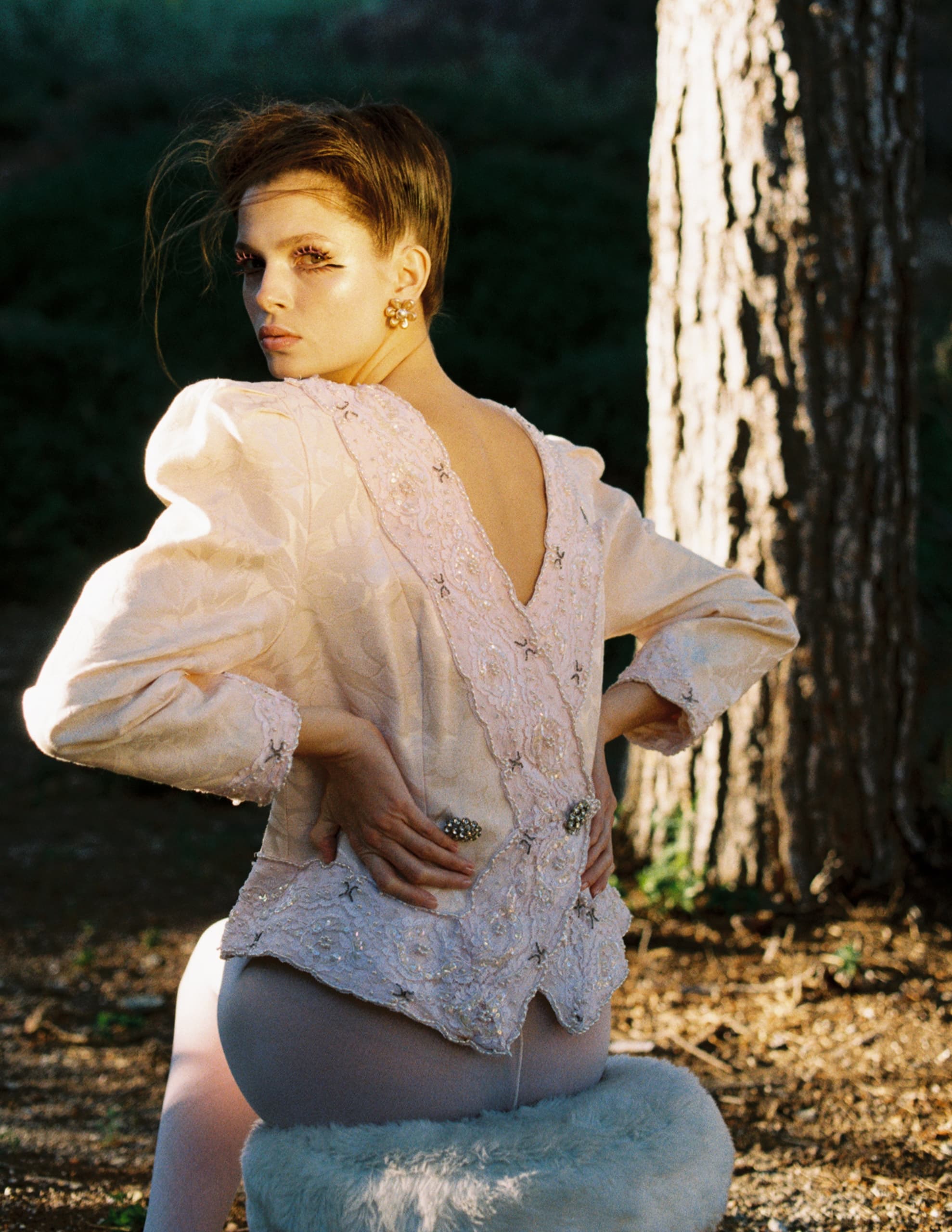 Woman in vintage lace jacket posing in sunlit forest setting.