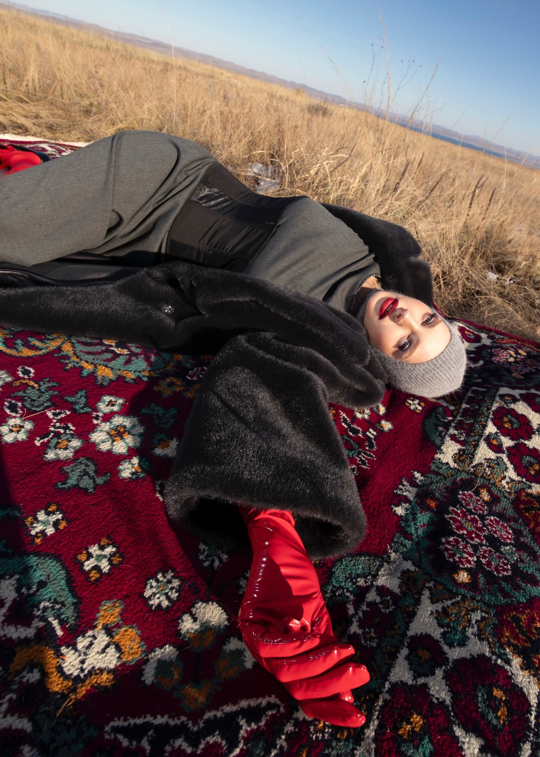 Fashionable woman in fur coat with red gloves lying on patterned rug outdoors.