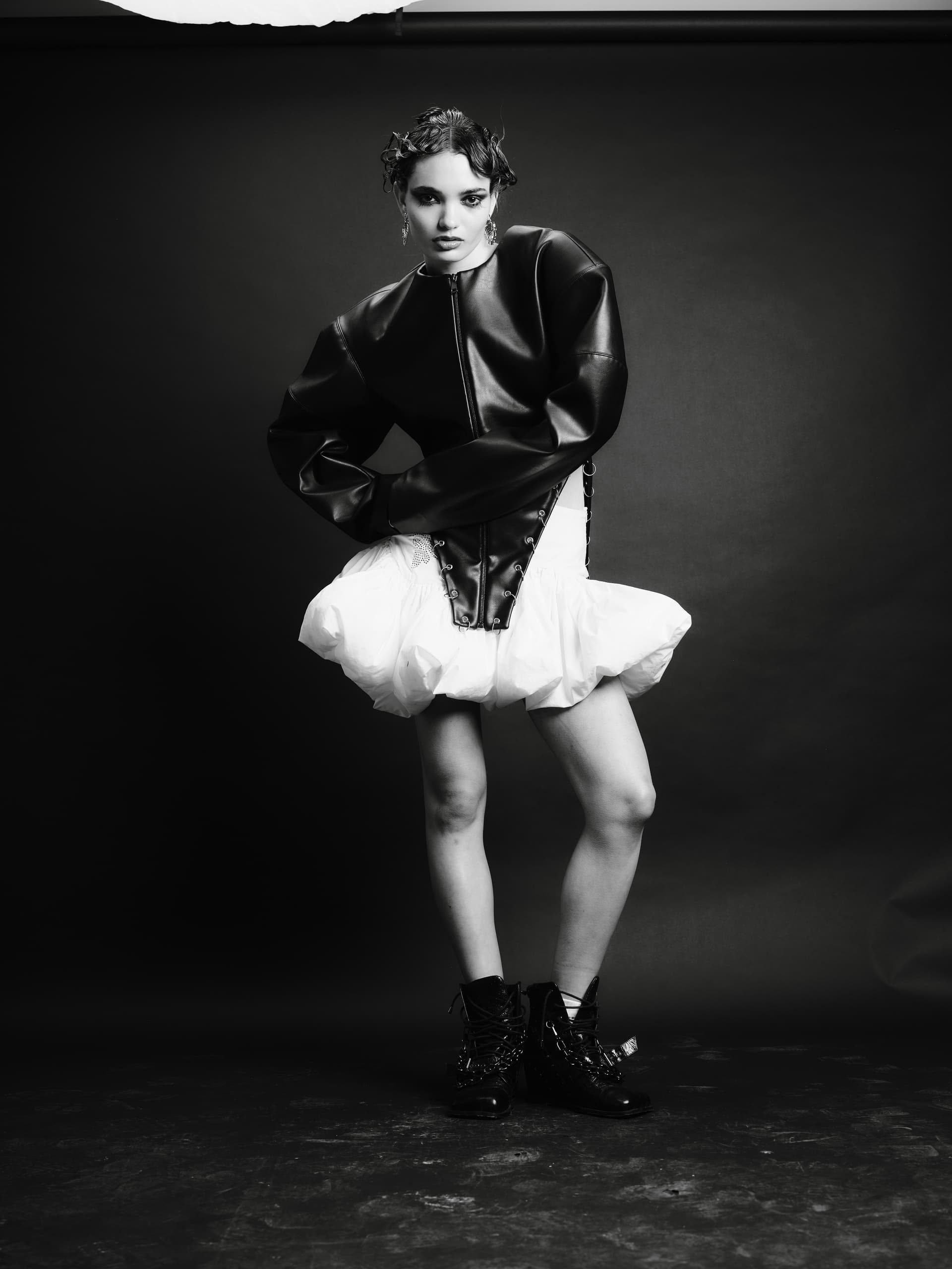 Fashion model in edgy black and white outfit with leather and ruffles, dark backdrop.