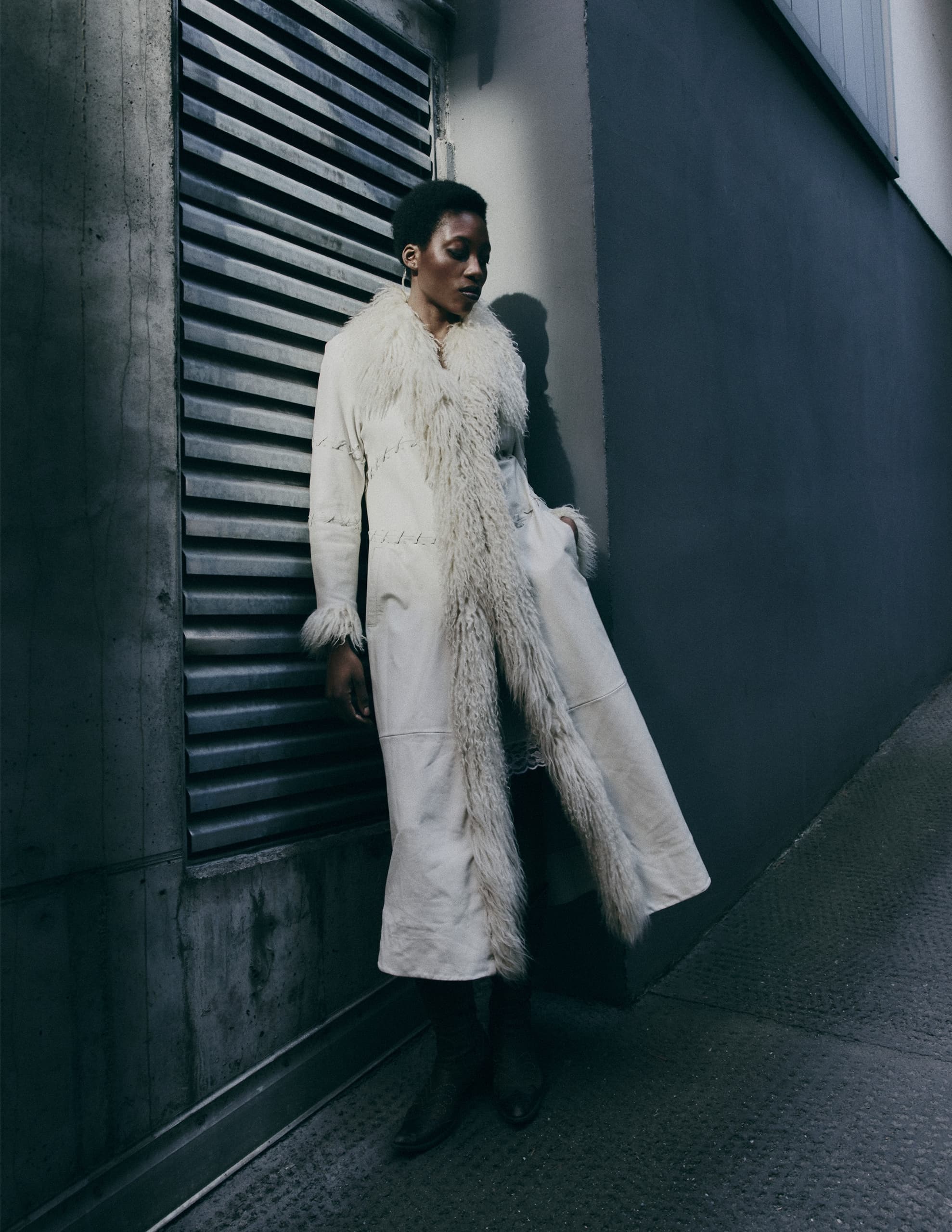 Stylish woman in trendy long white faux fur coat leaning against urban wall.