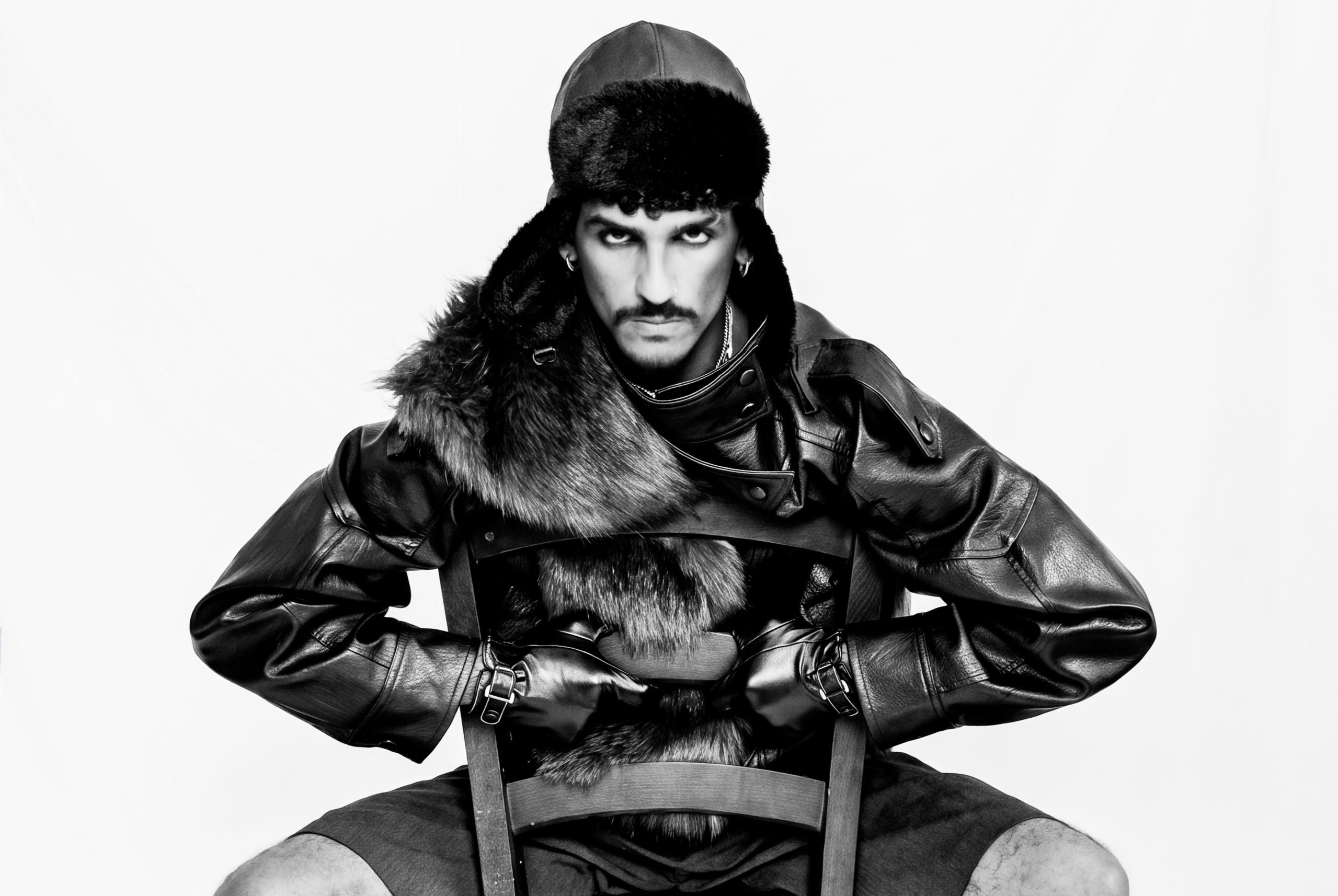 Man in edgy leather fashion and fur hat seated against a white background.