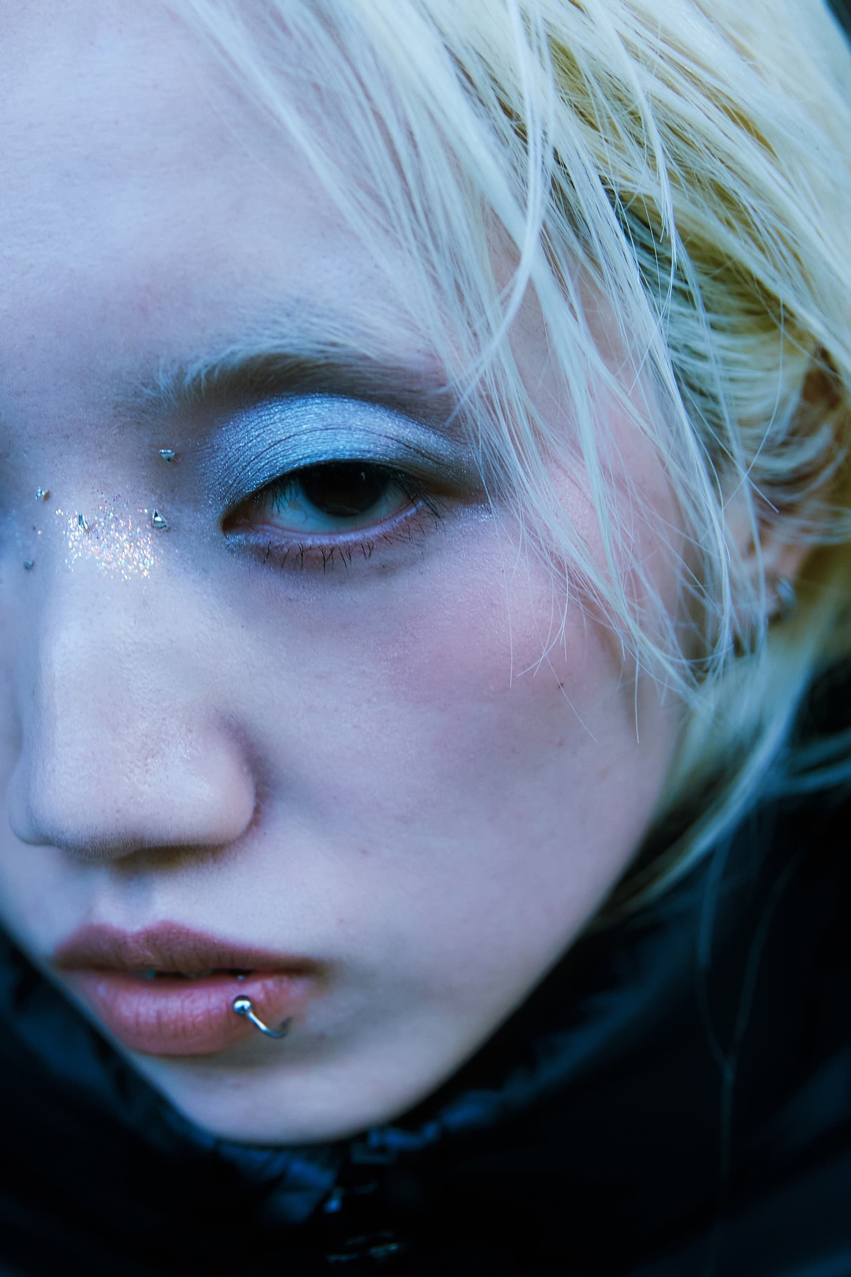 Close-up of person with blue eyeshadow, glitter, and lip piercing.