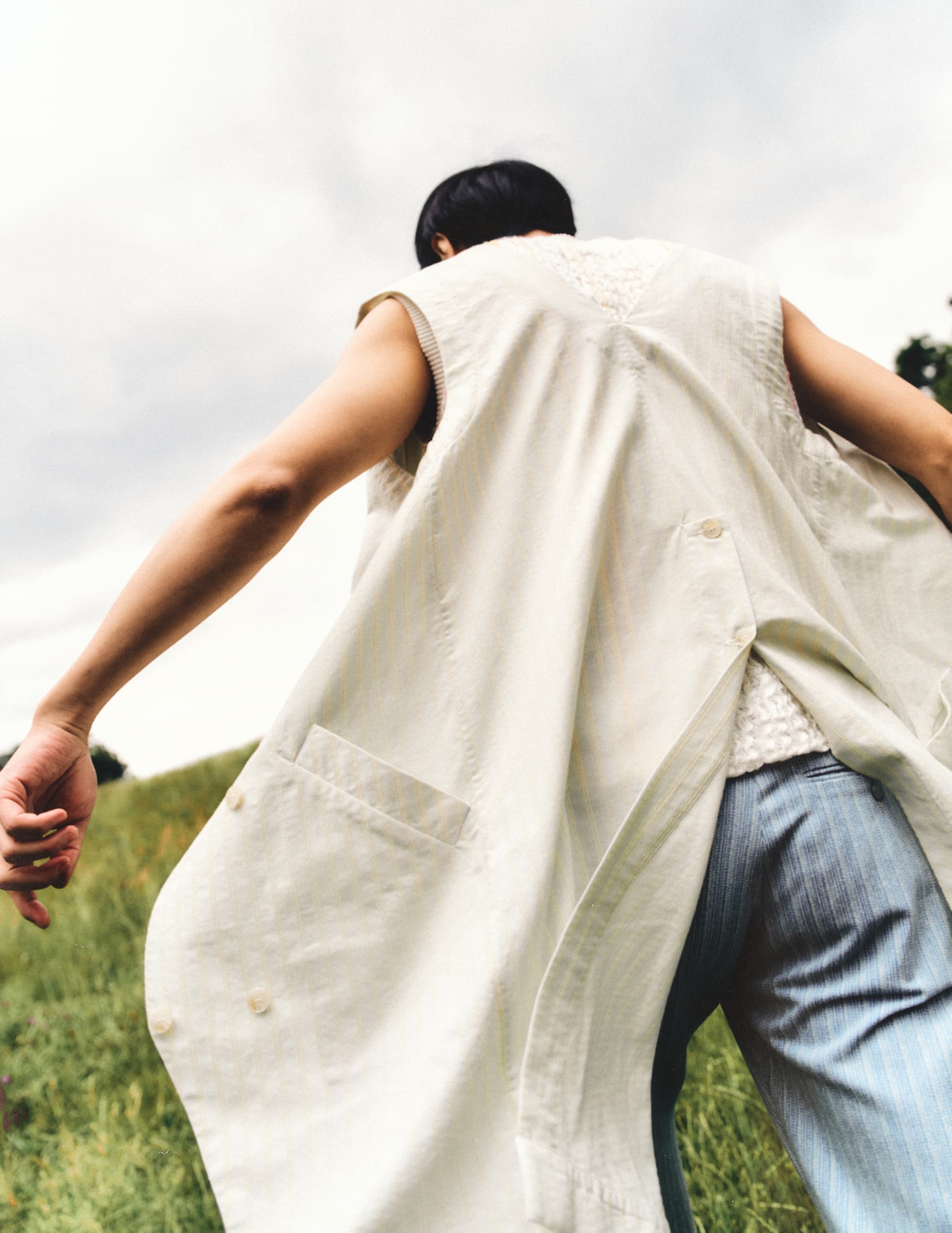 Person in flowy white vest walking through grassy field under cloudy sky.