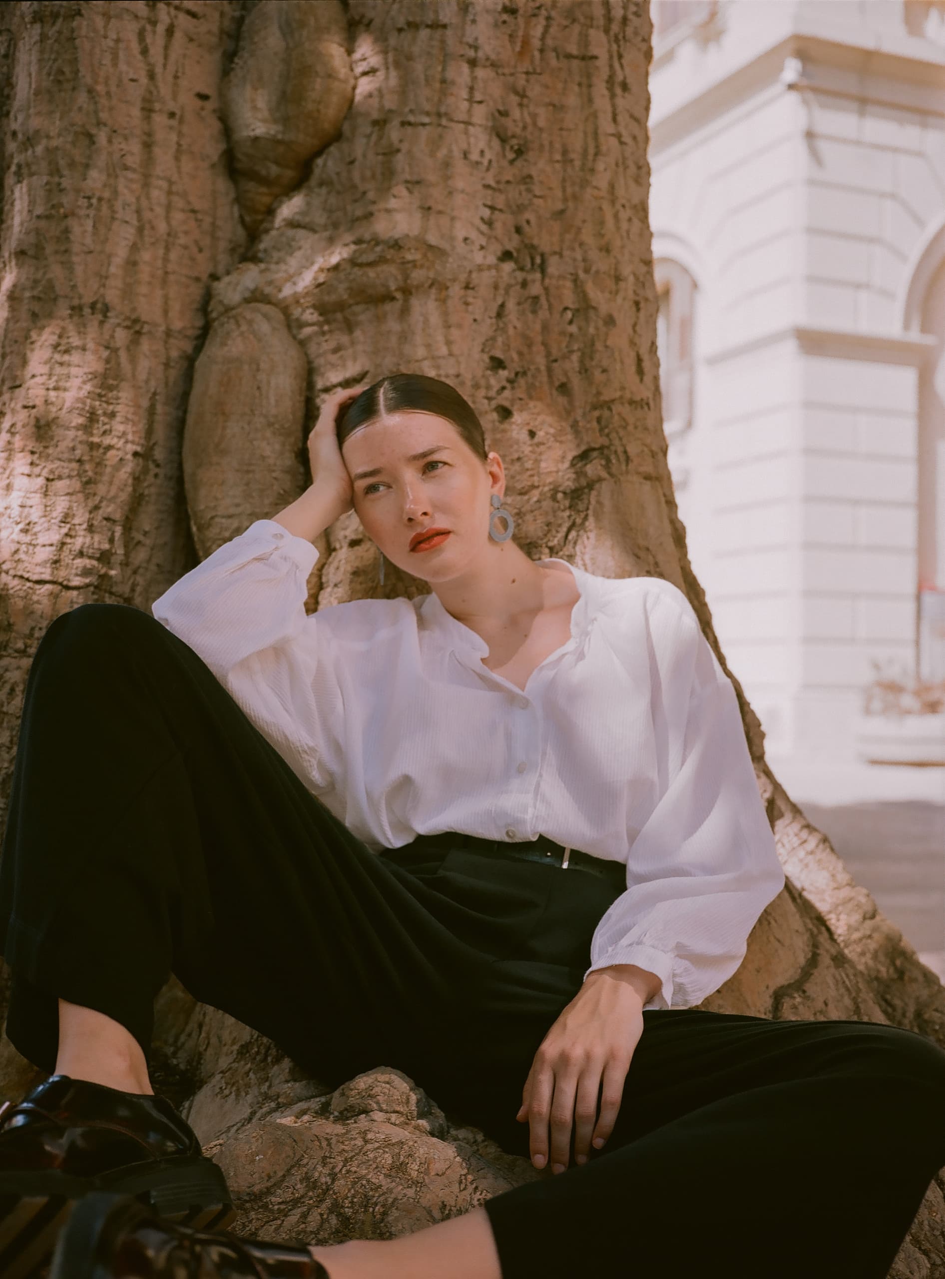 Woman in white blouse sitting by a tree in a thoughtful pose.