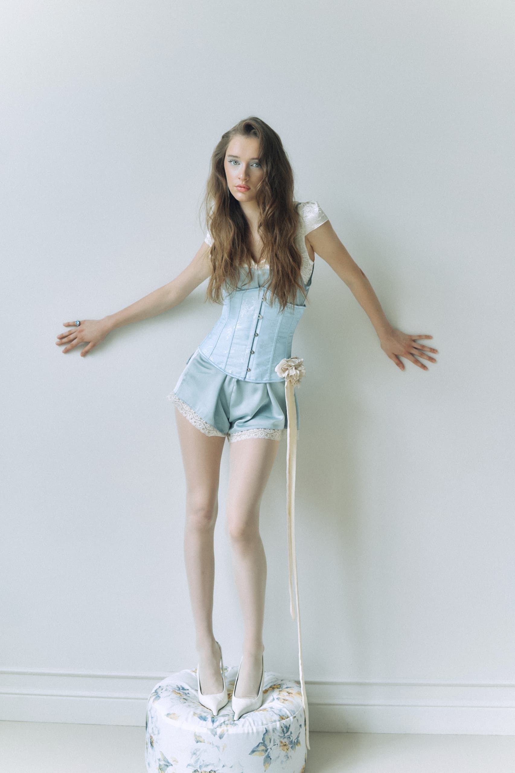 Woman in vintage blue corset and shorts standing on floral stool.