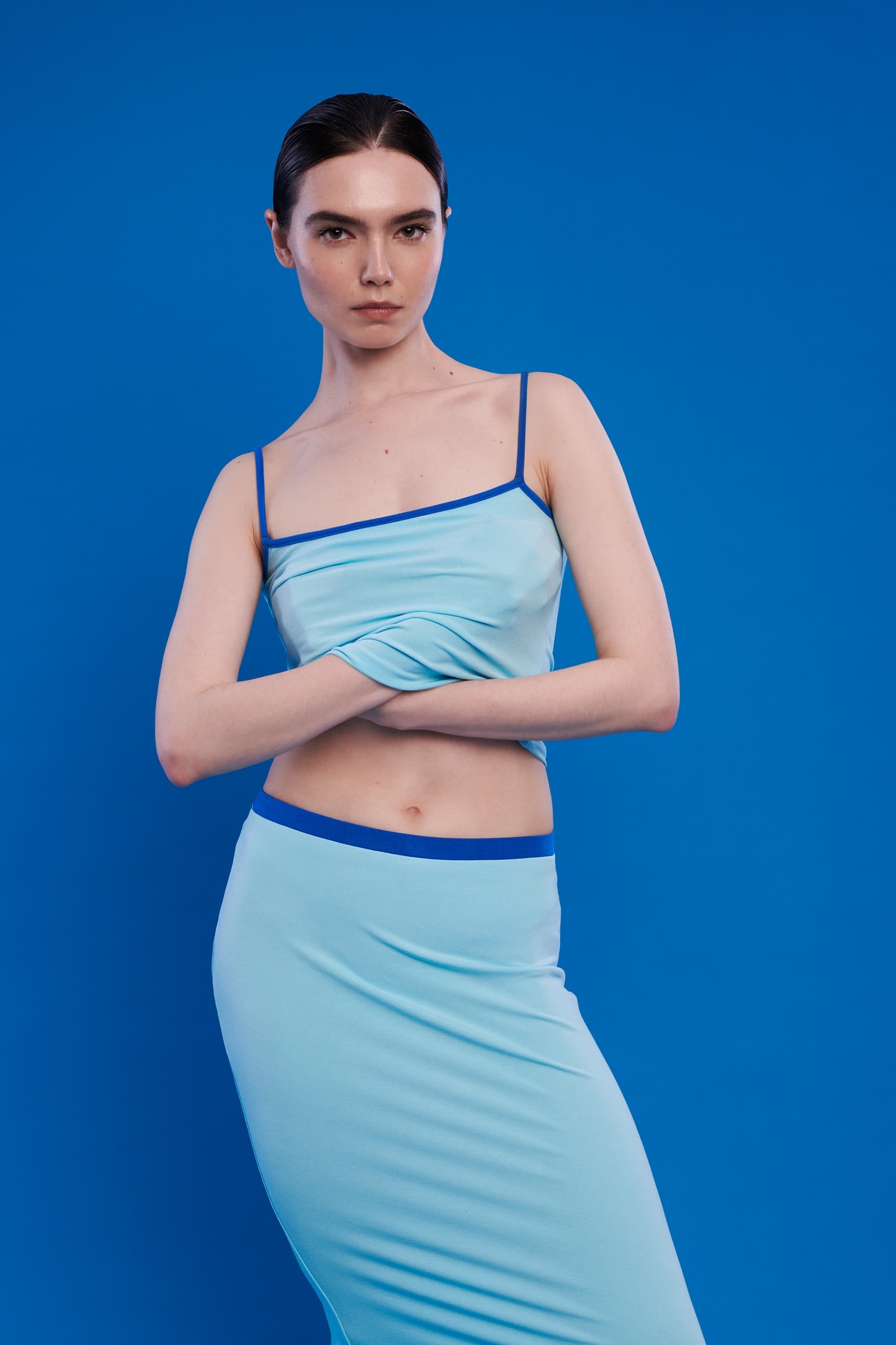 Woman in light blue outfit poses confidently with arms crossed, blue background.