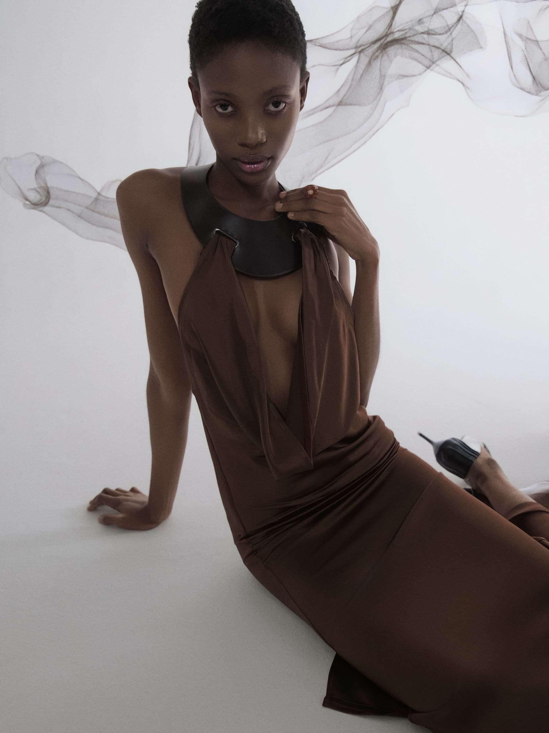 Model in elegant brown dress with flowing fabric background, fashion photography.