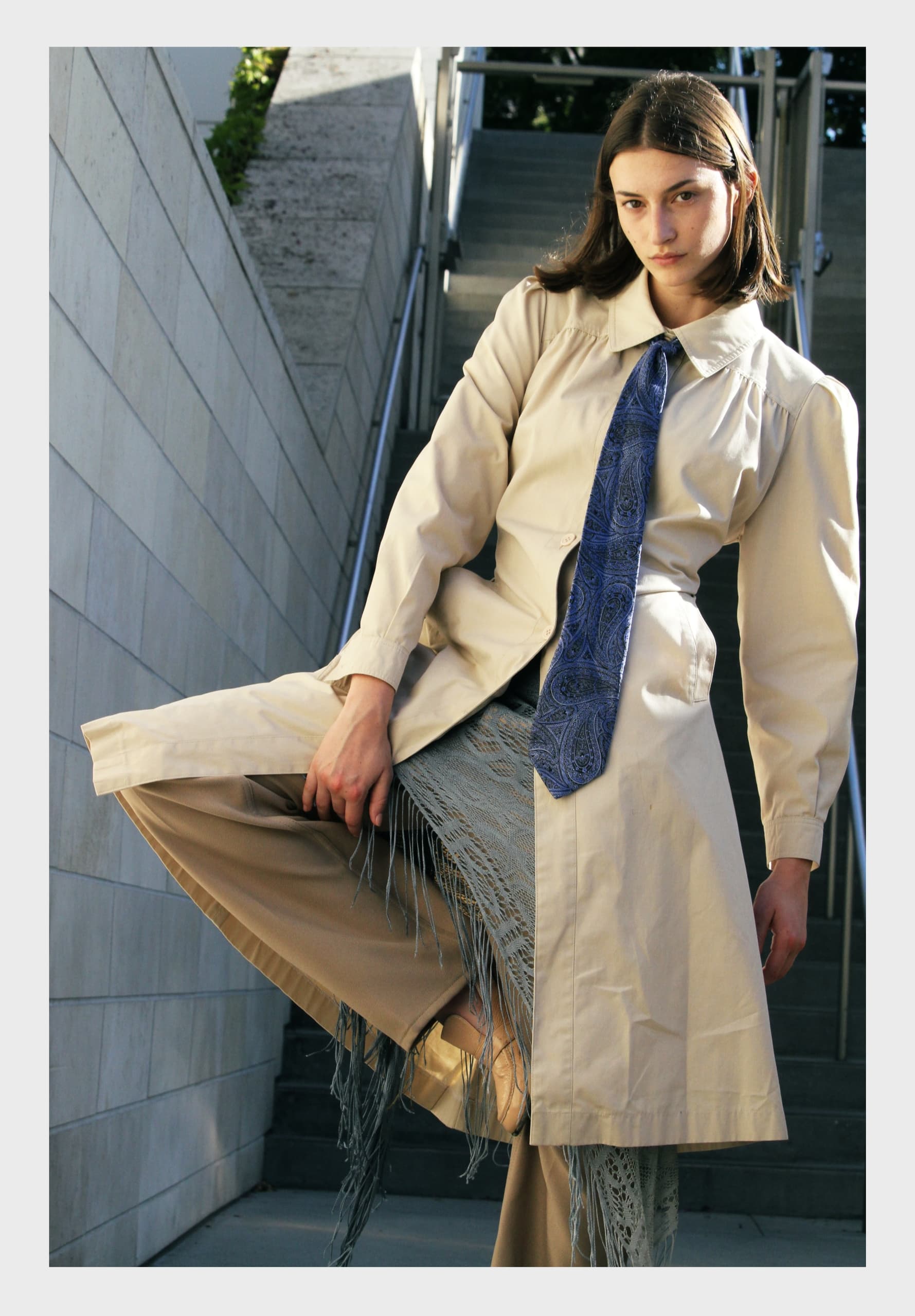 Fashionable woman in beige outfit and blue tie striking a stylish pose on stairs.