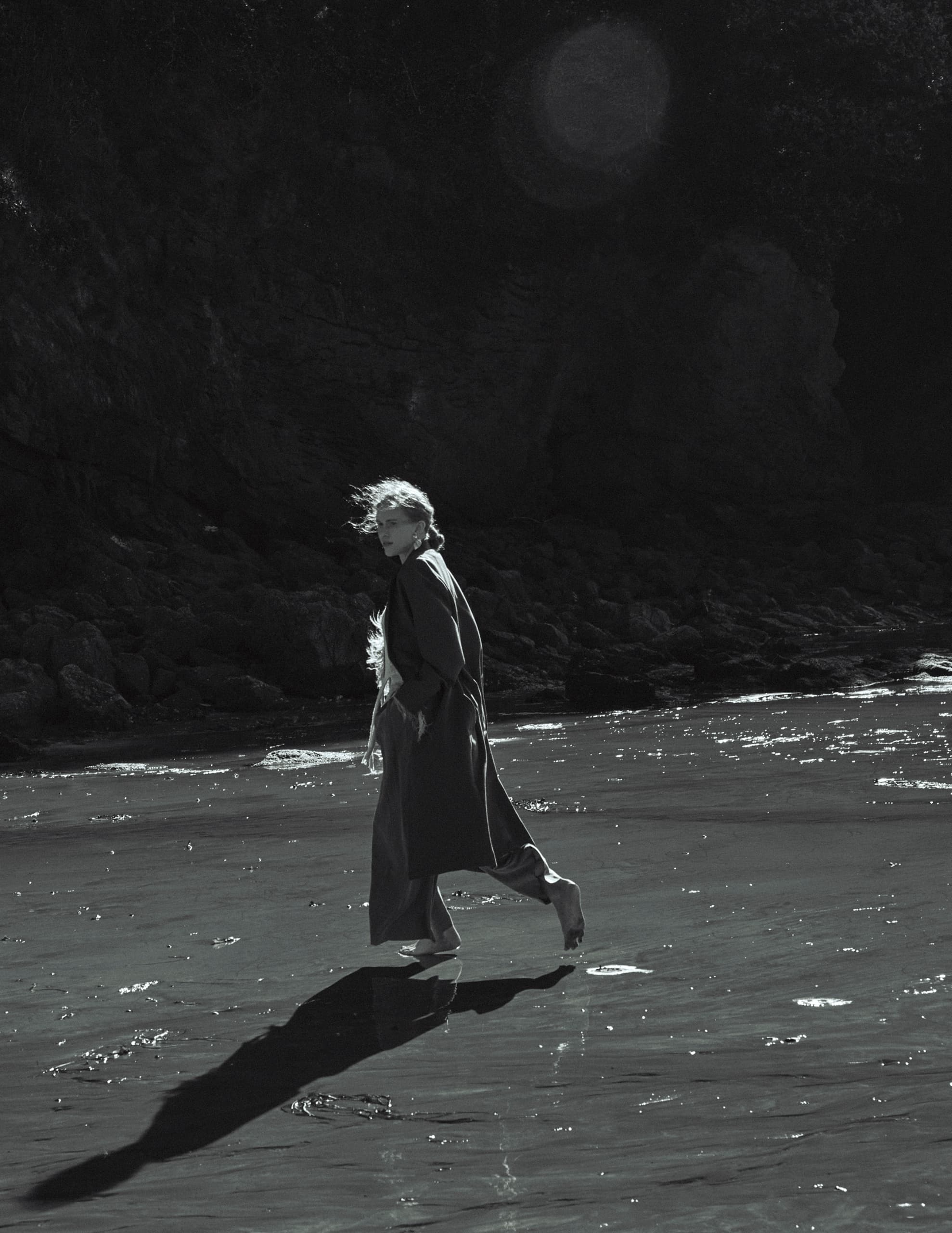 Person walking on seaside in dramatic lighting, casting long shadow on wet sand.