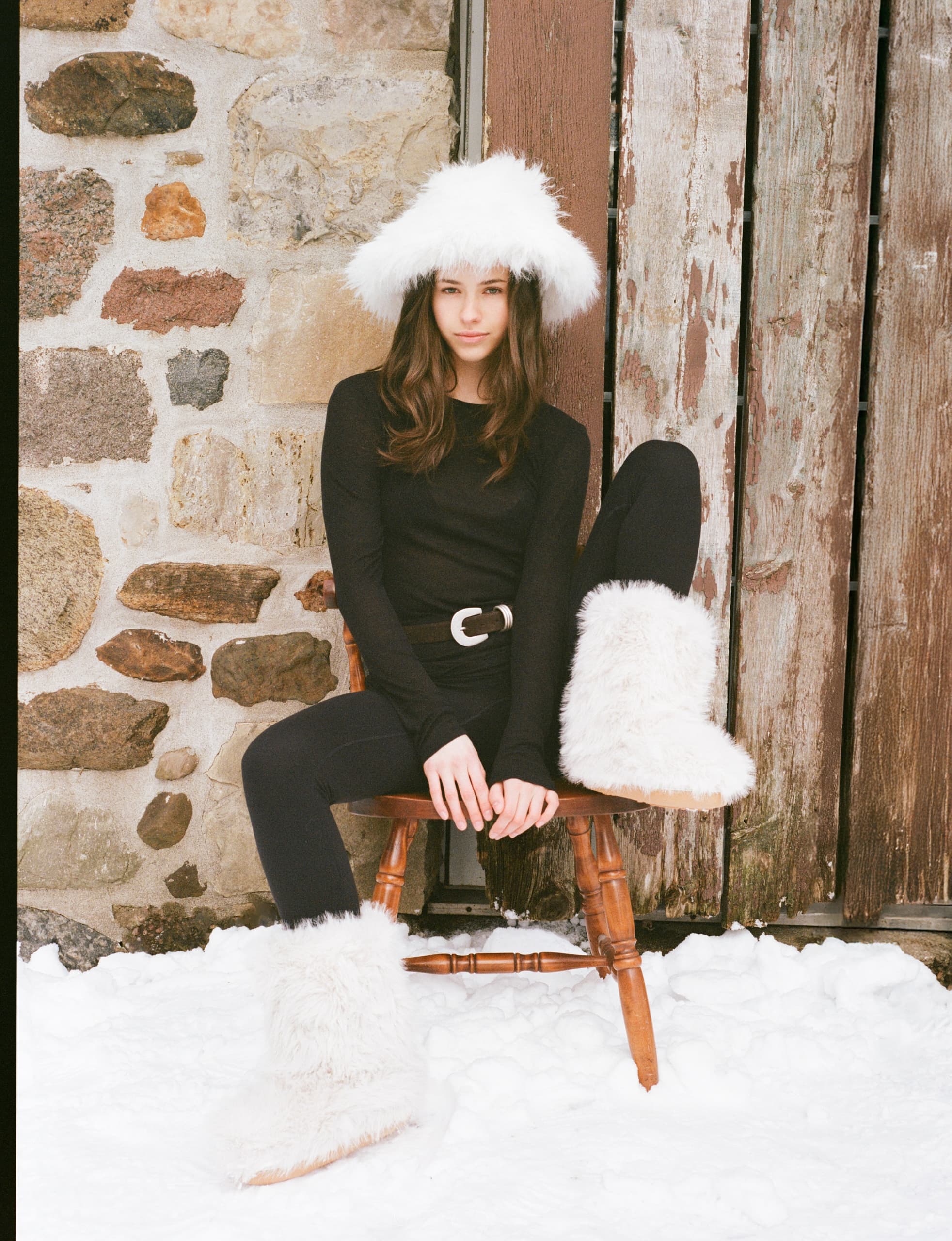 Woman in winter outfit with fur hat and boots, seated in snowy rustic setting.