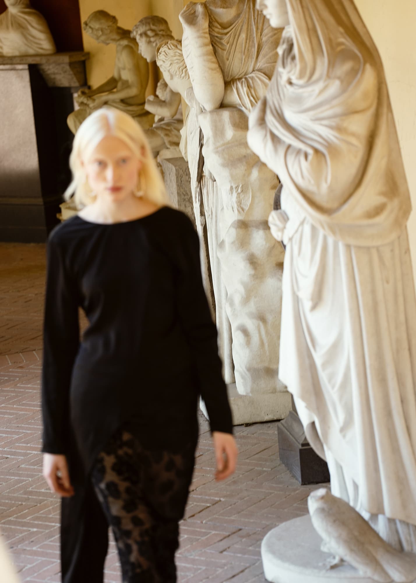 Model in black outfit walking past classical sculptures.