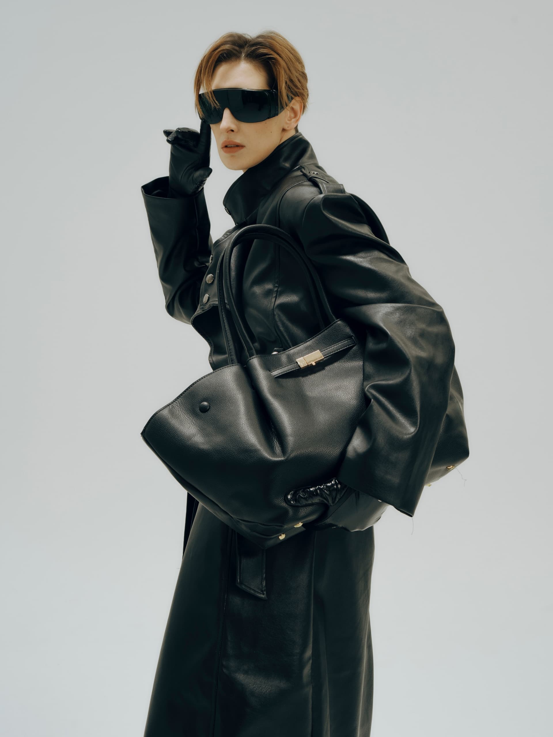 Stylish person in black leather coat and sunglasses, holding a large leather handbag.