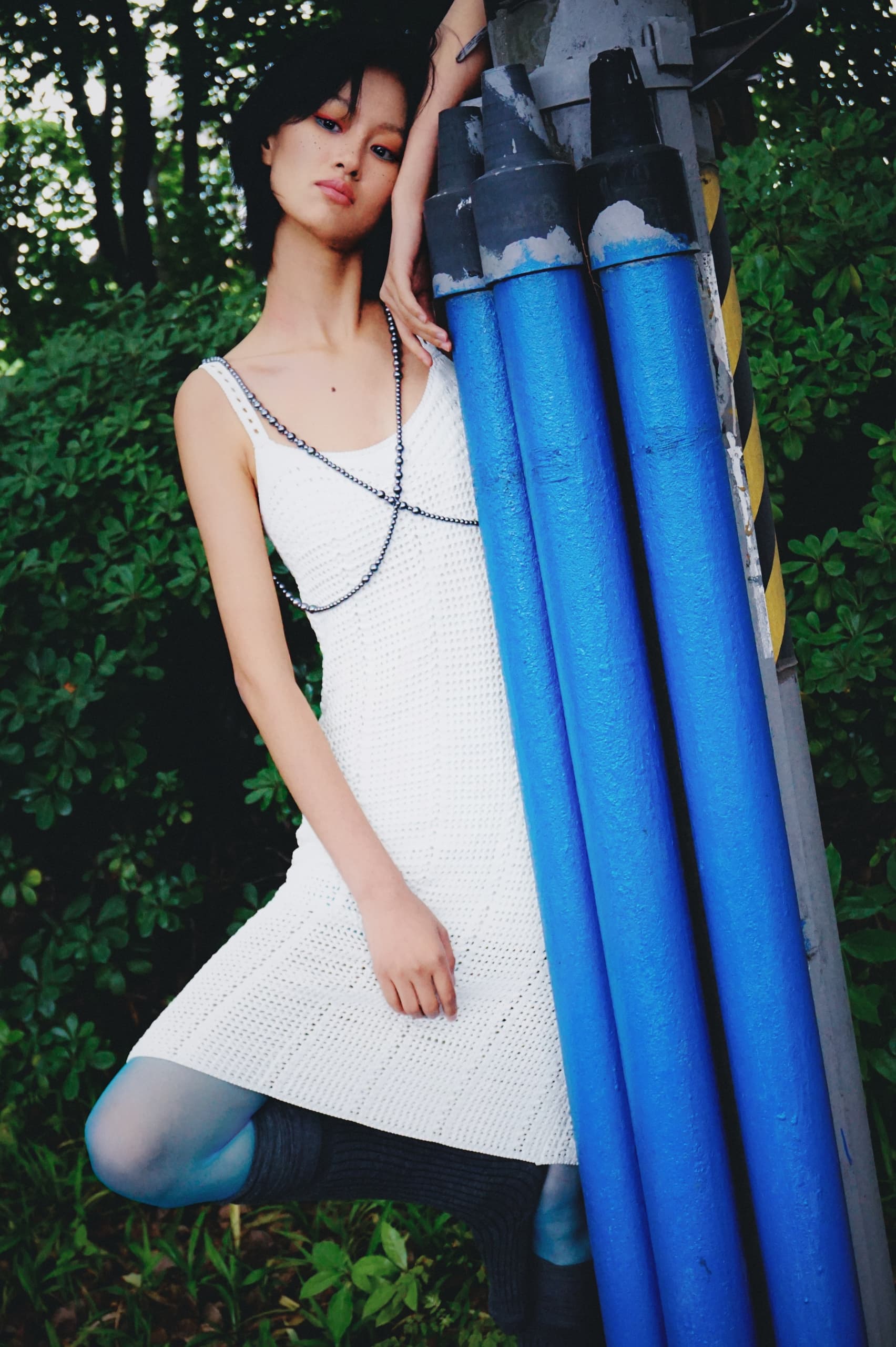 Fashionable woman in white dress leaning against blue pipes outdoors.
