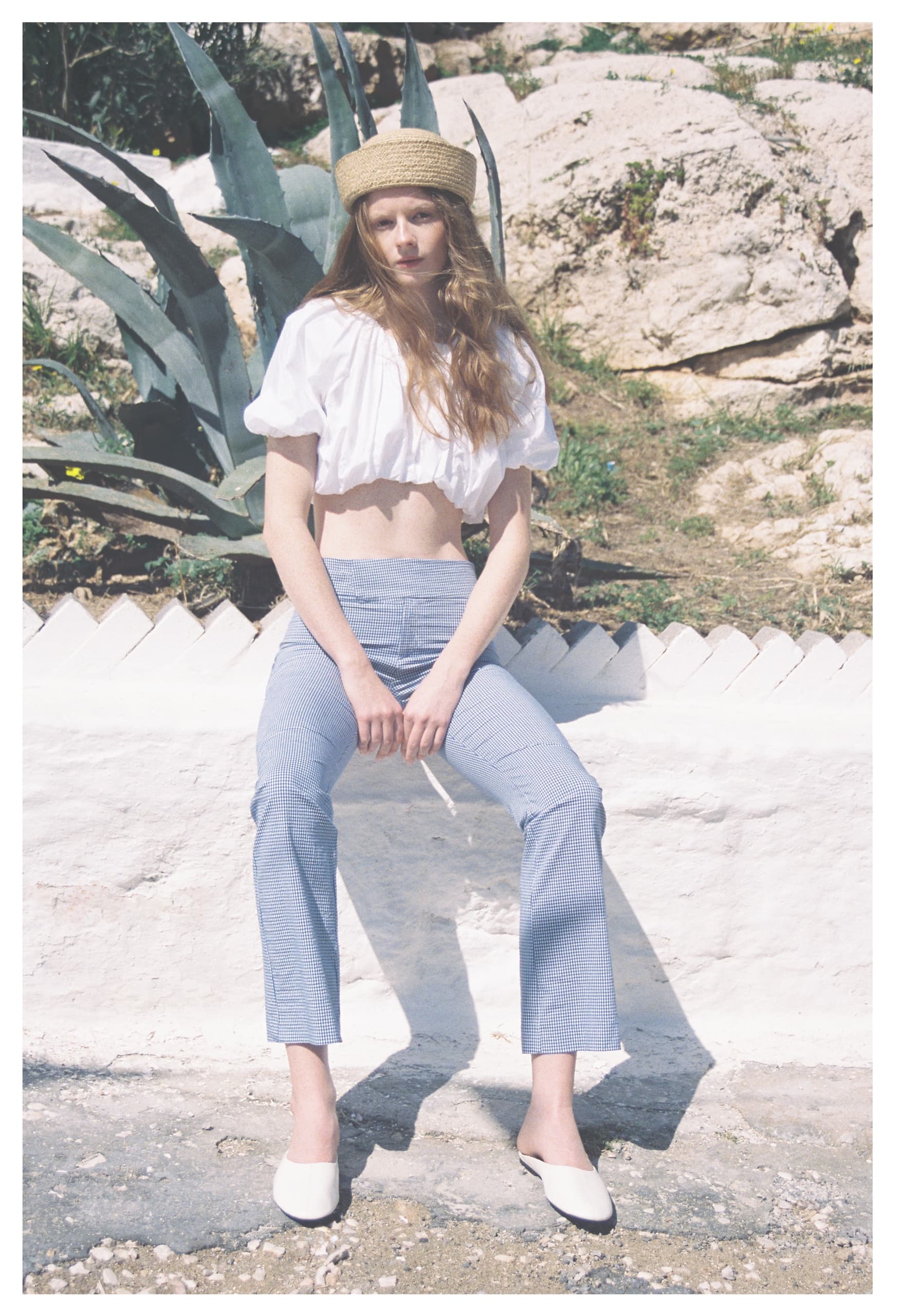 Woman in white top, blue pants, sitting outdoors with plants and rocks in the background.
