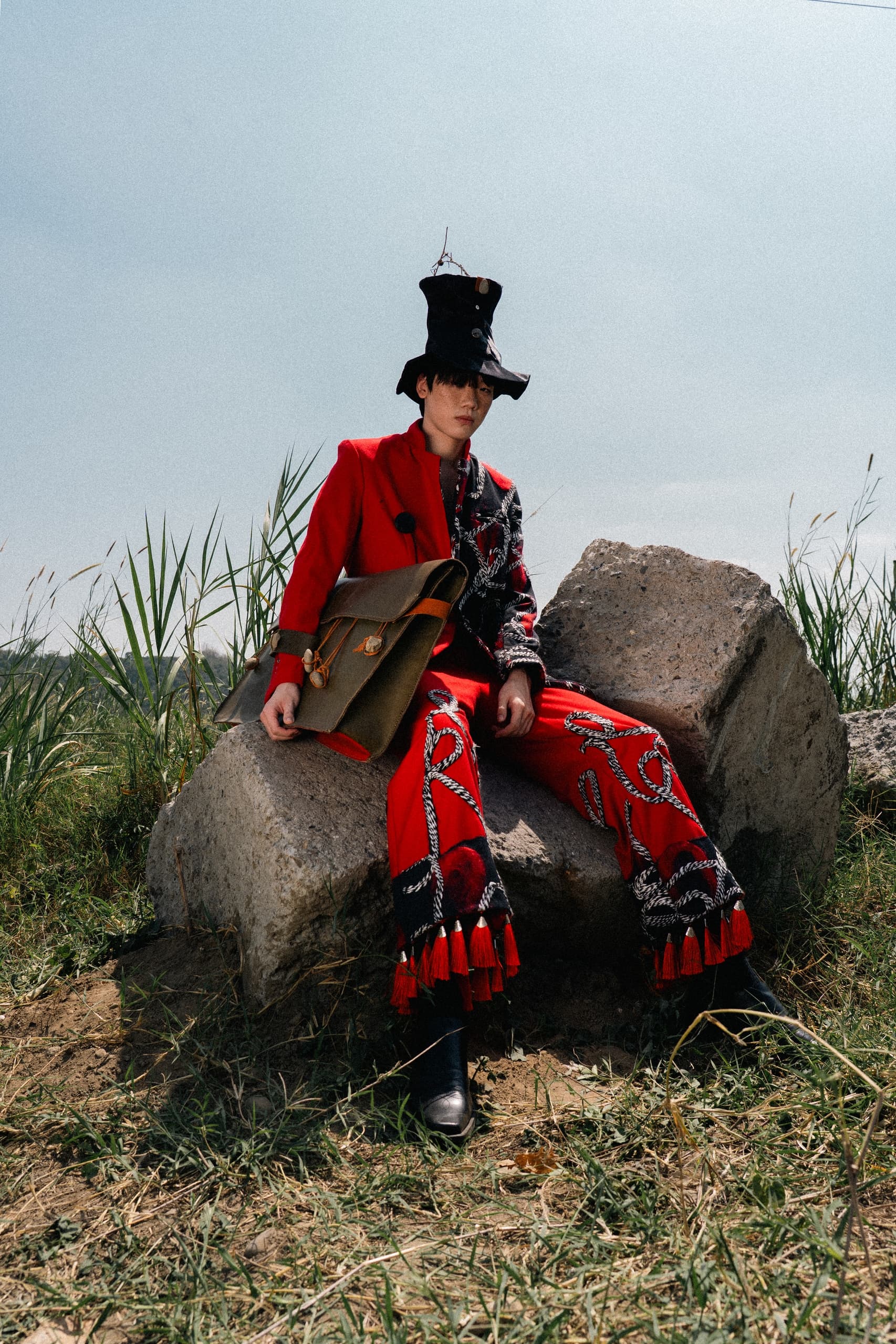 Person in avant-garde red outfit with unique hat sitting on rocks outdoors.
