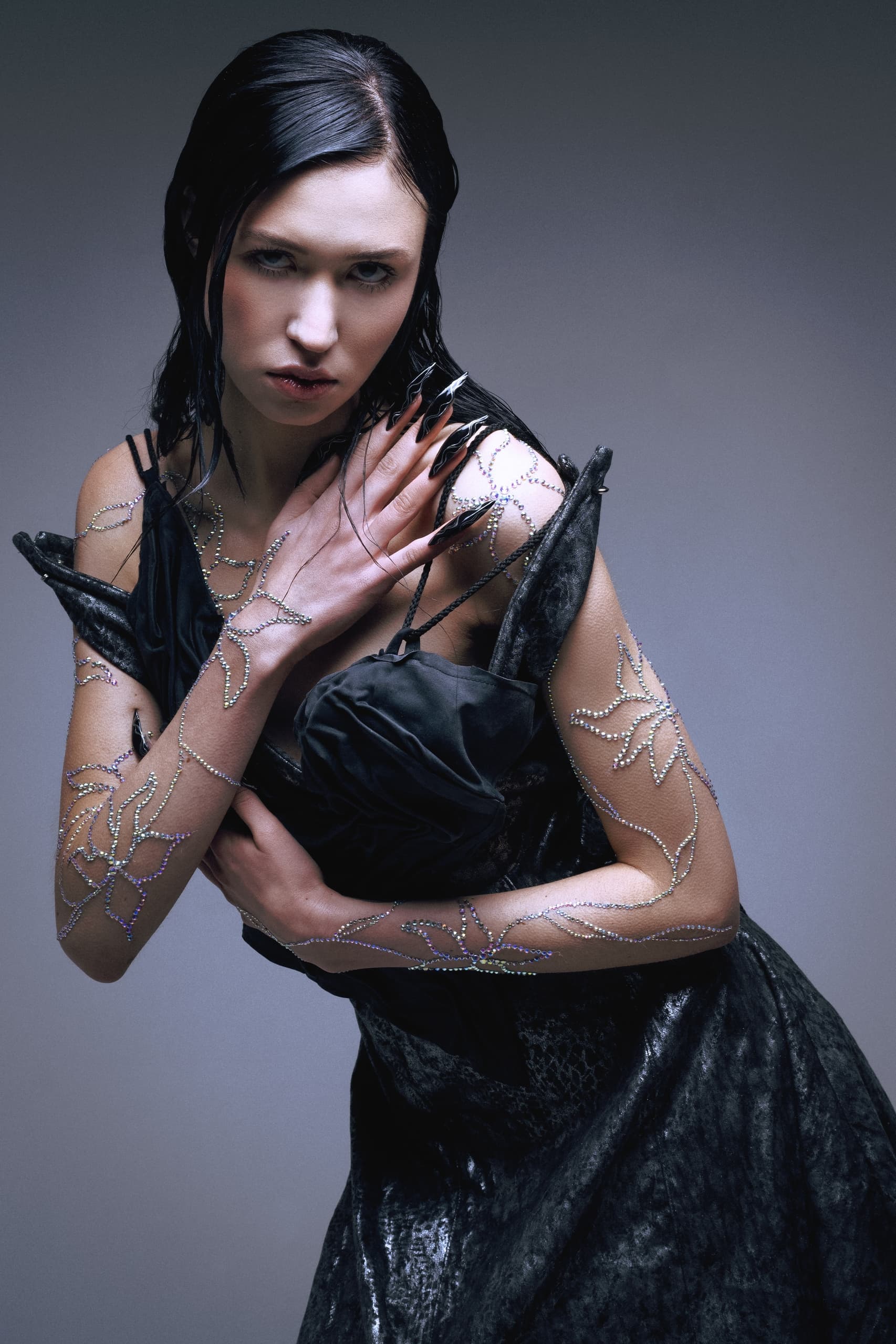 Gothic fashion model with dark nails and rhinestone arm designs in dramatic pose.