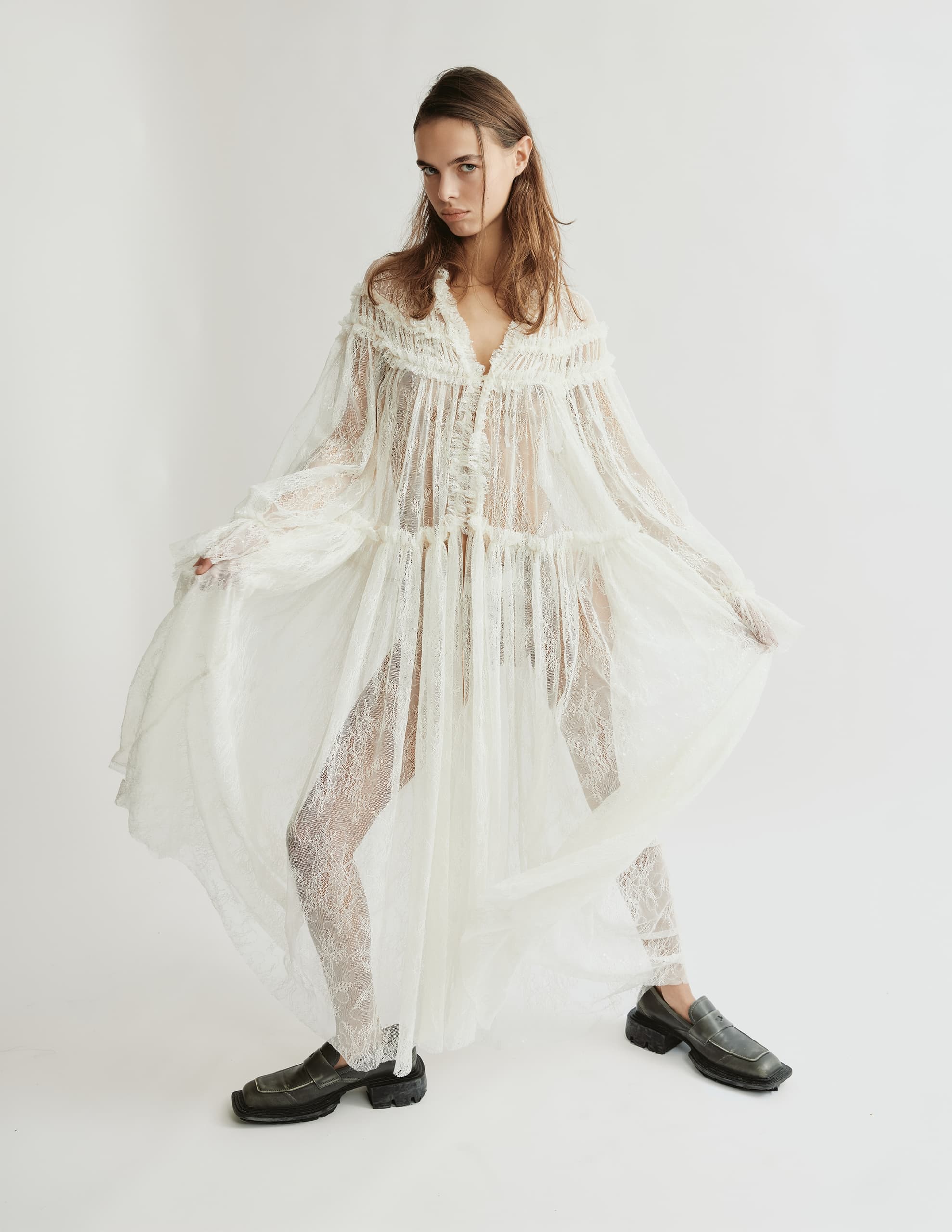 Model in sheer lace dress and chunky shoes on white background.