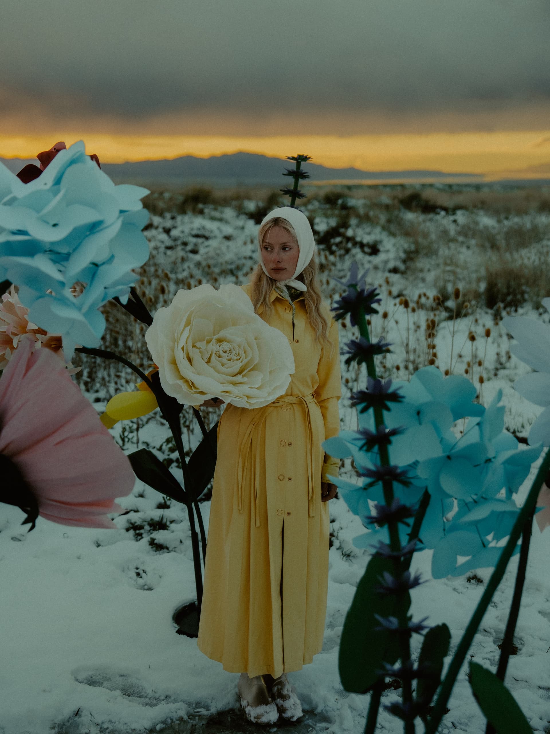 Woman in yellow dress stands among giant flowers in a snowy landscape at sunset.