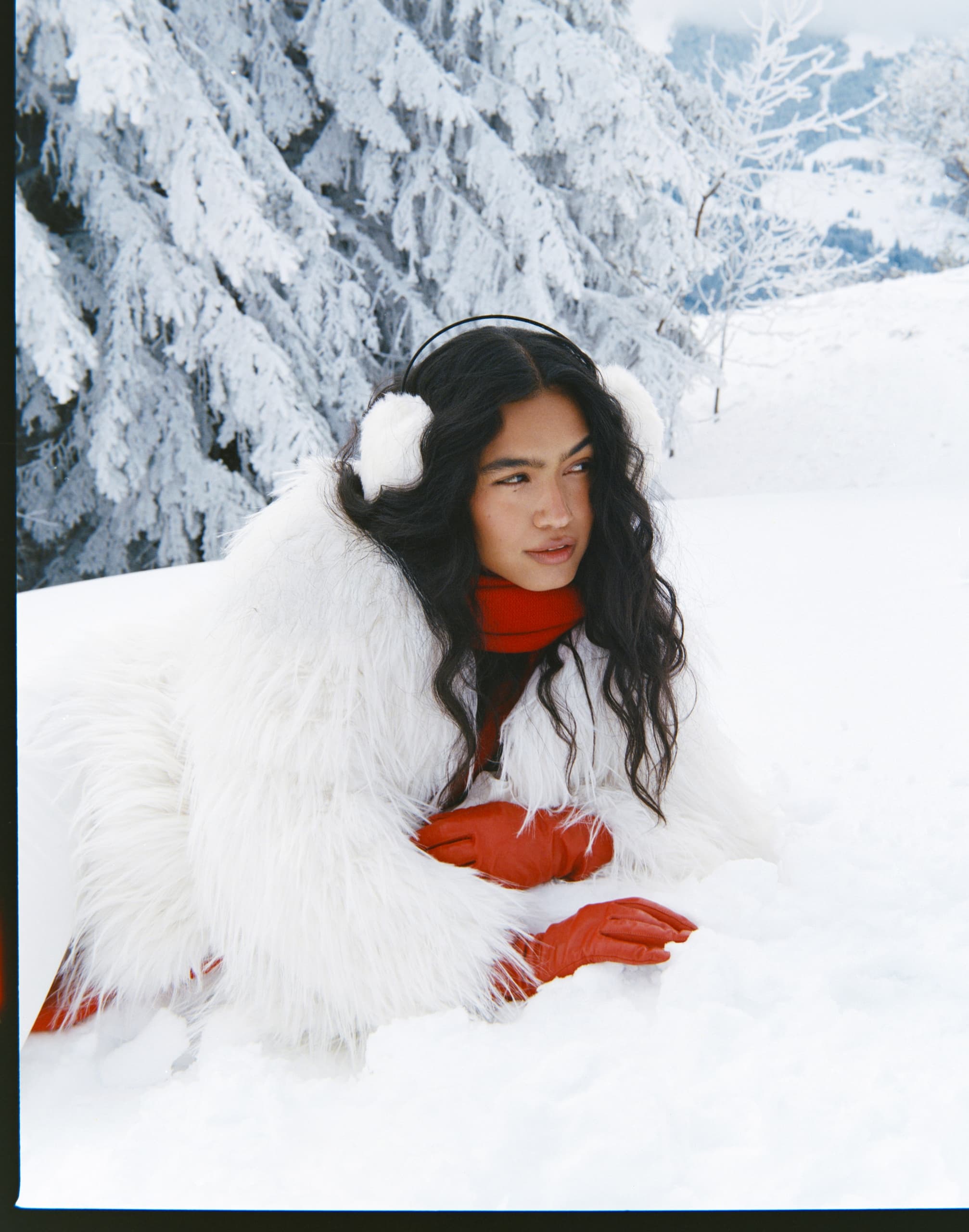 Woman in snowy forest wearing cozy winter attire, fluffy earmuffs, and red gloves.