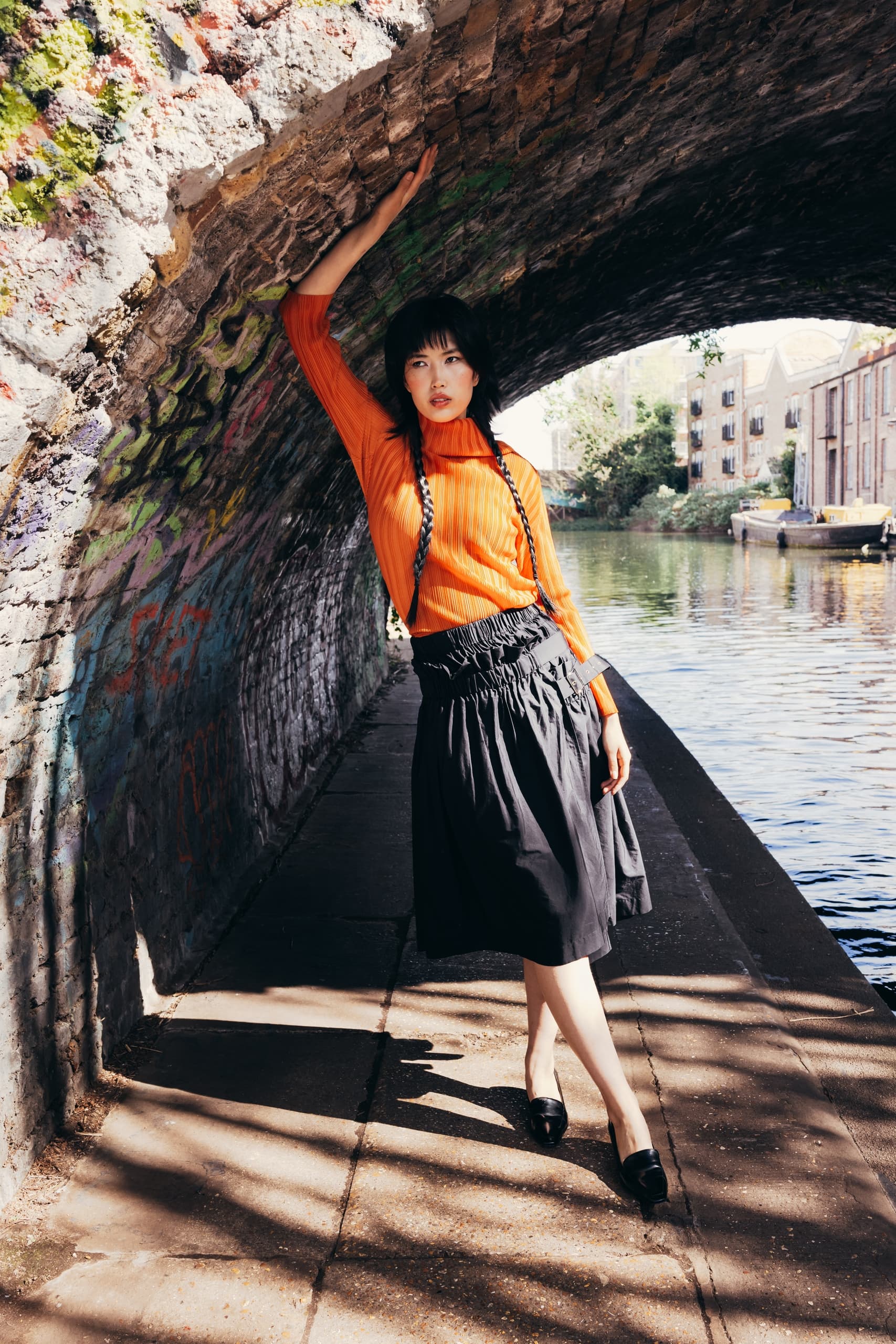 Model posing stylishly under a brick arch by a canal on a sunny day.