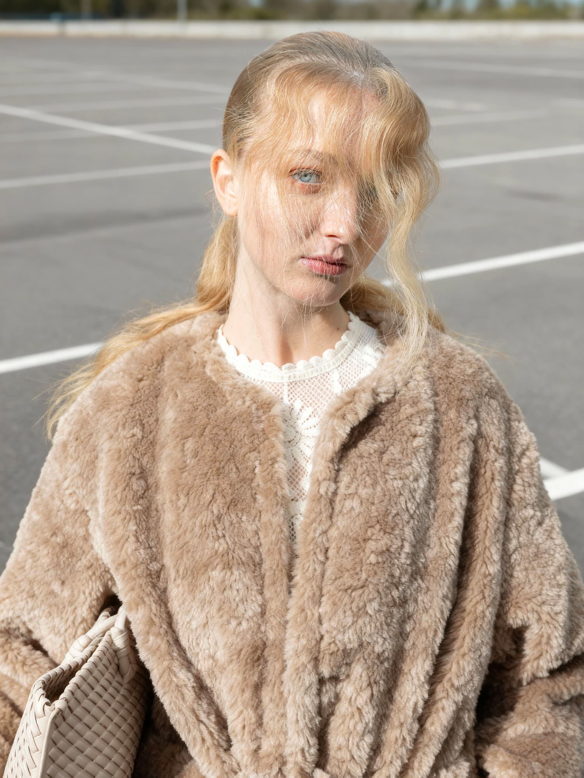 Person in fur coat standing in an empty parking lot, holding a woven clutch bag.