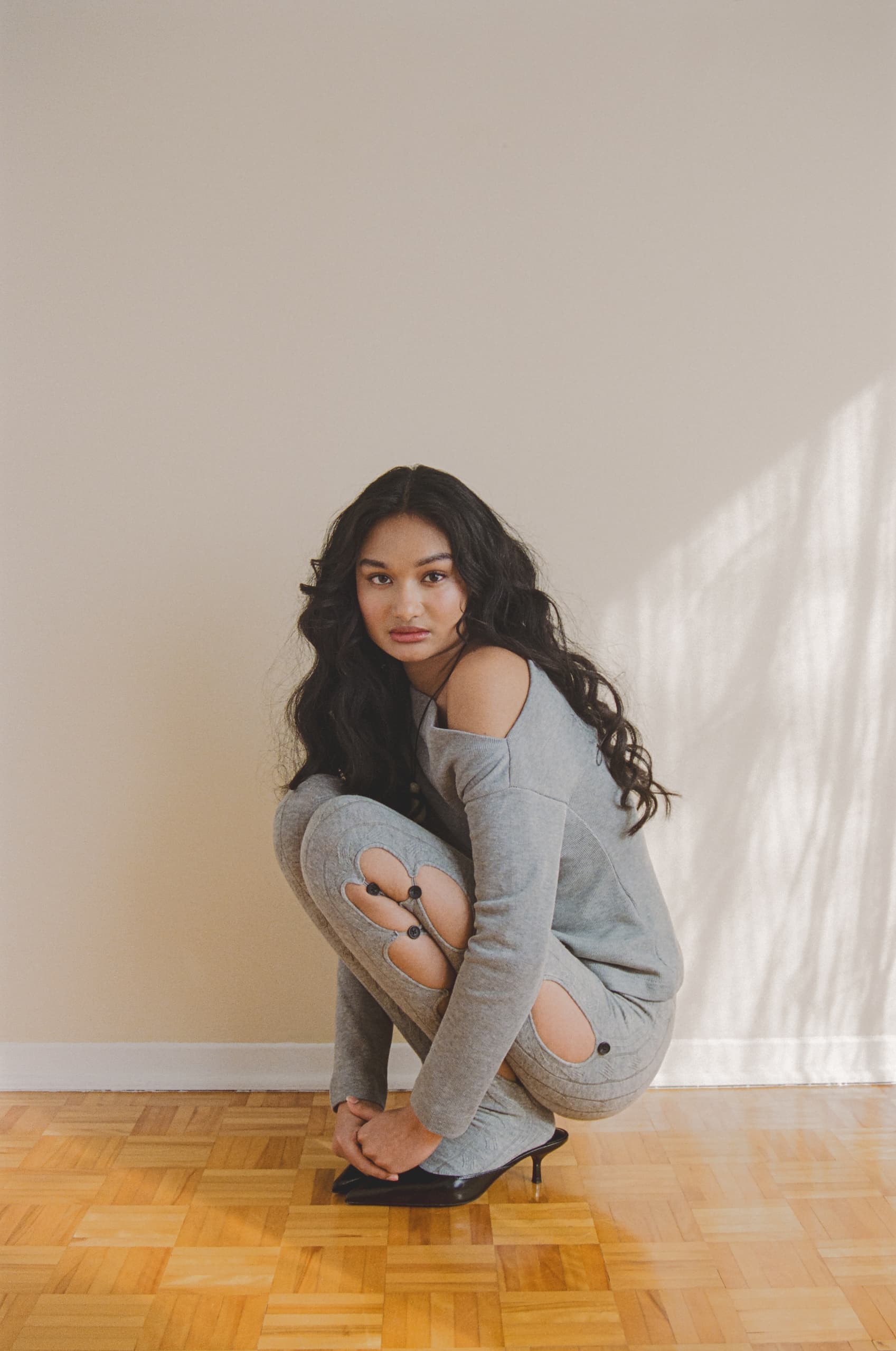 Woman in gray outfit with cutouts crouches on wood floor, sunlight on the wall.