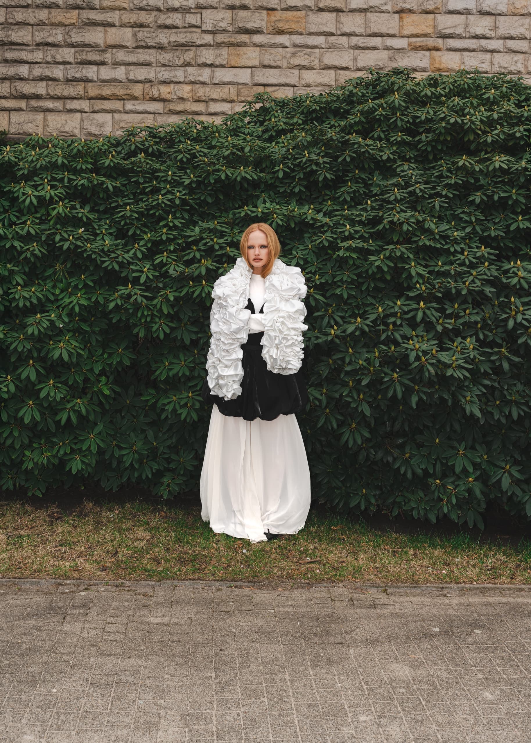 Woman in elegant black and white outfit stands against lush greenery background.