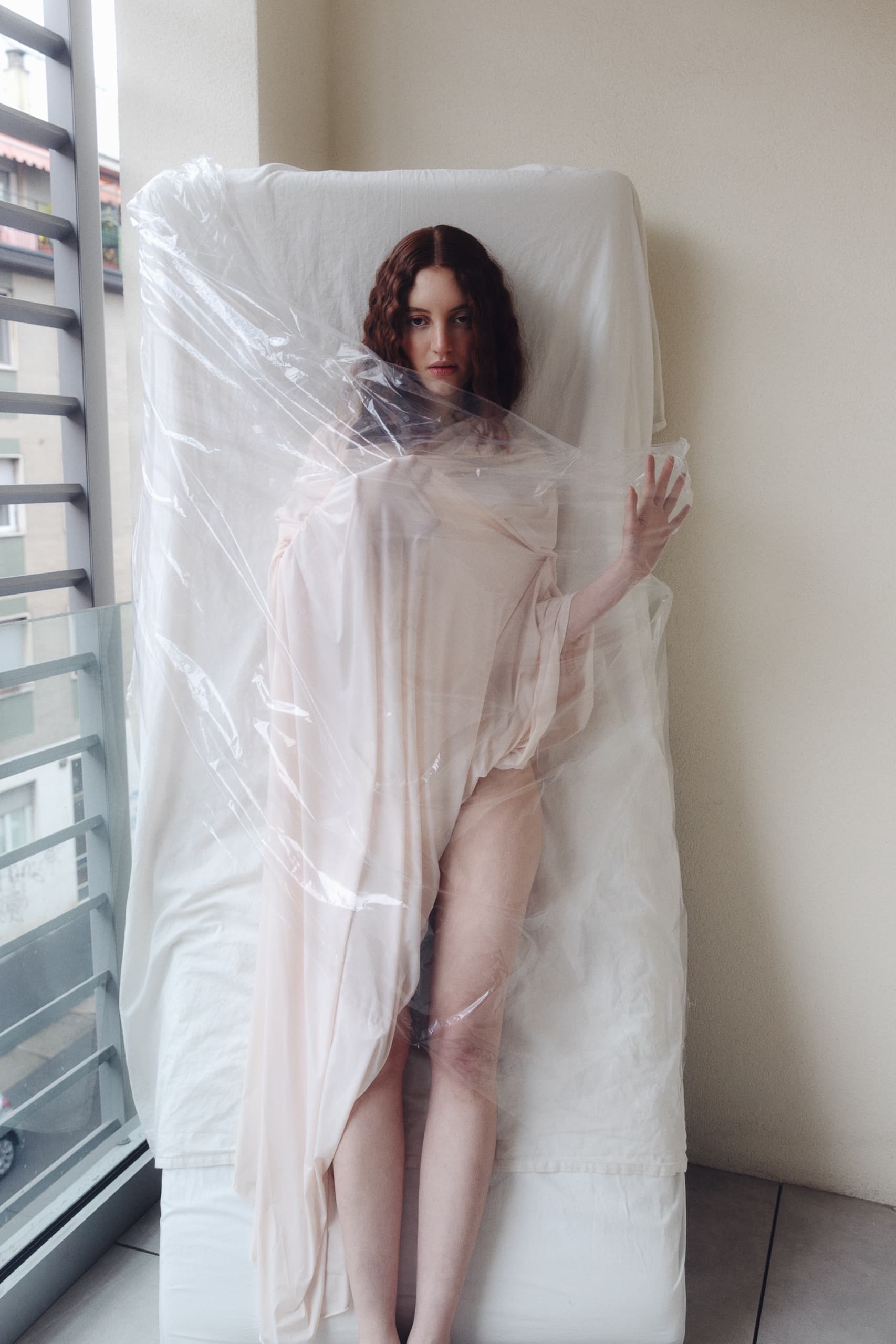 Woman wrapped in sheer fabric and plastic on a bed, artistic indoor portrait.