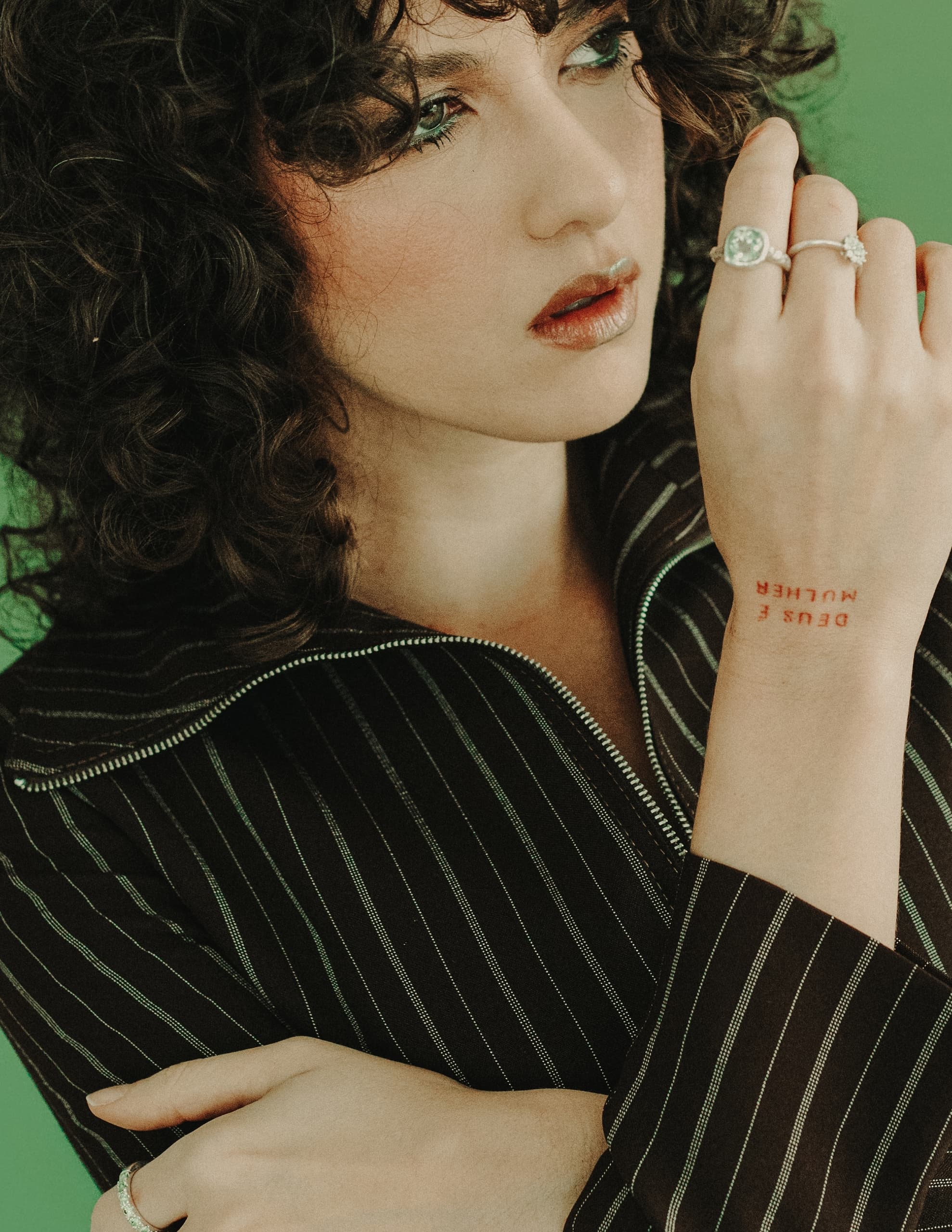 Curly-haired woman with striped top and rings, thoughtful expression, green background.