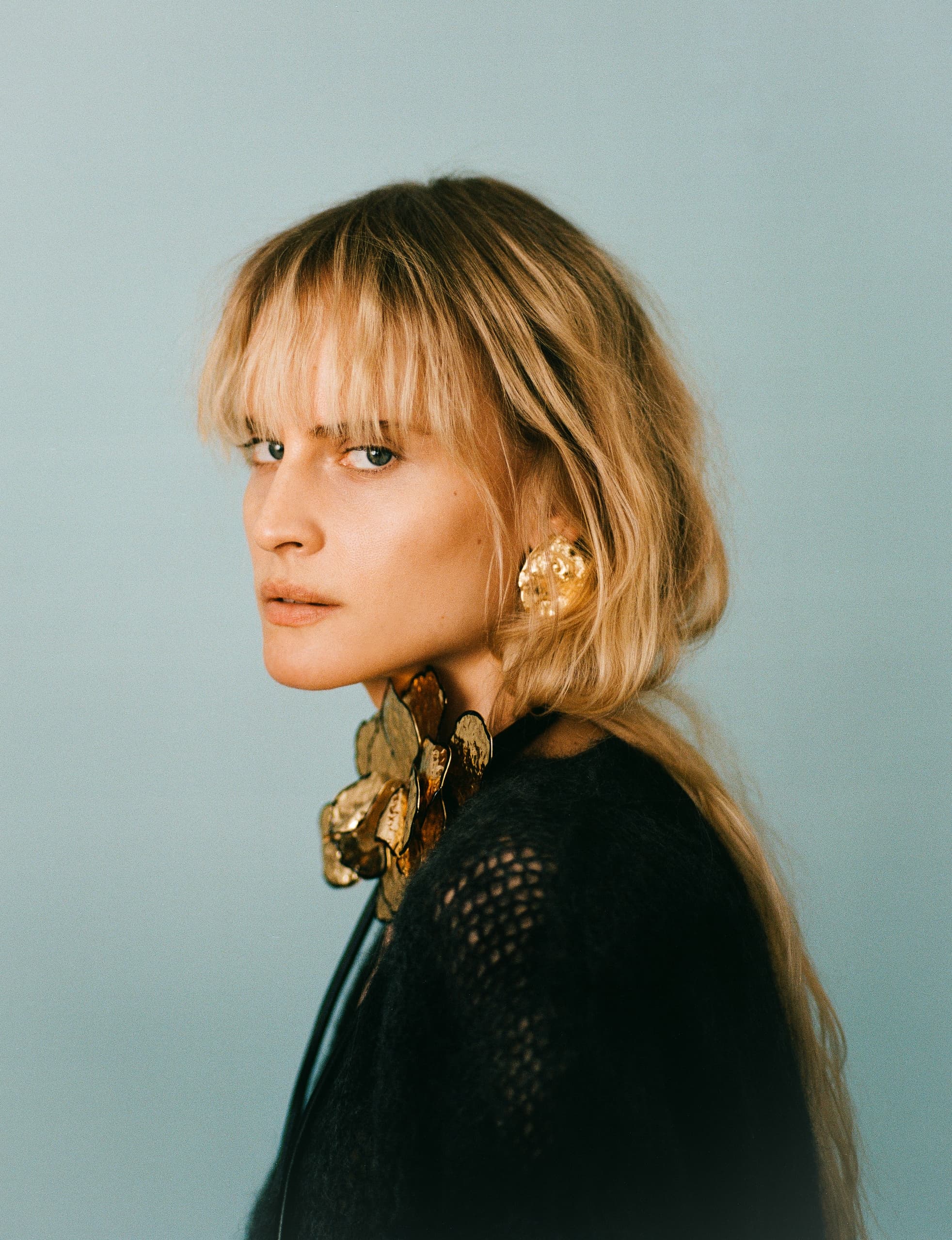 Elegant woman with blonde hair wearing gold jewelry against a light blue background.