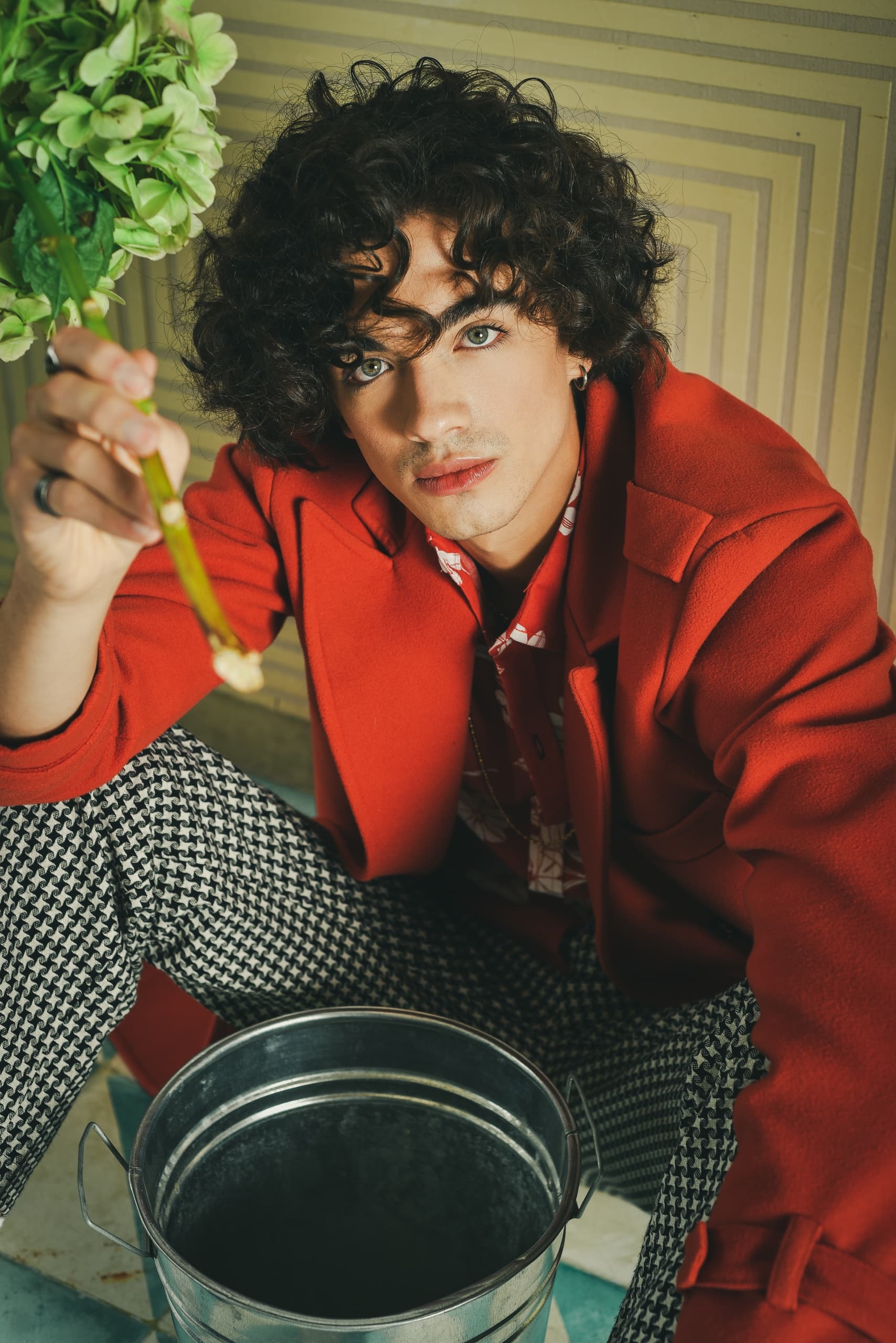 Person in red coat holding chopsticks, sitting by a metal bucket indoors.