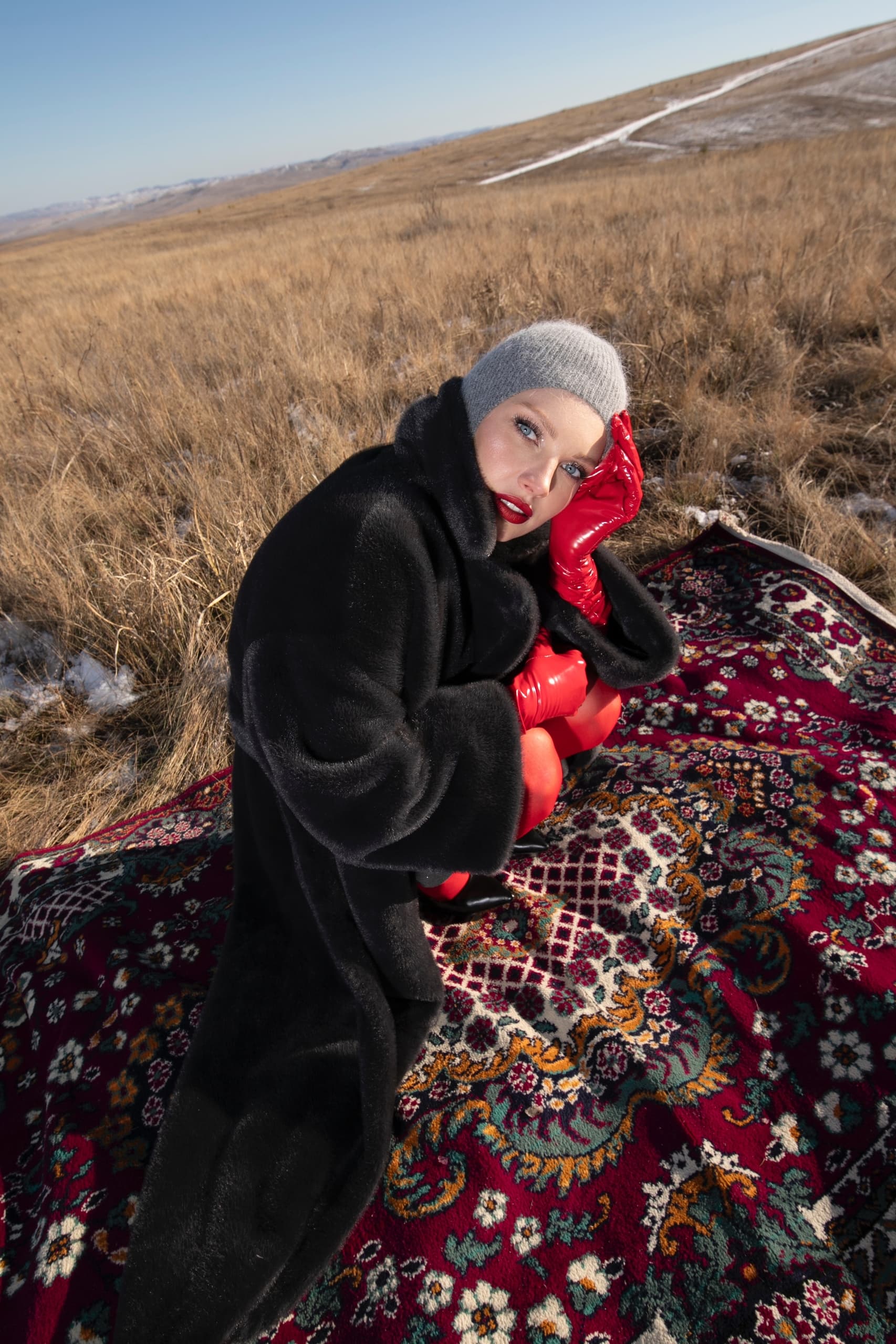 Person in winter coat on patterned blanket in field, wearing red gloves and gray beanie.