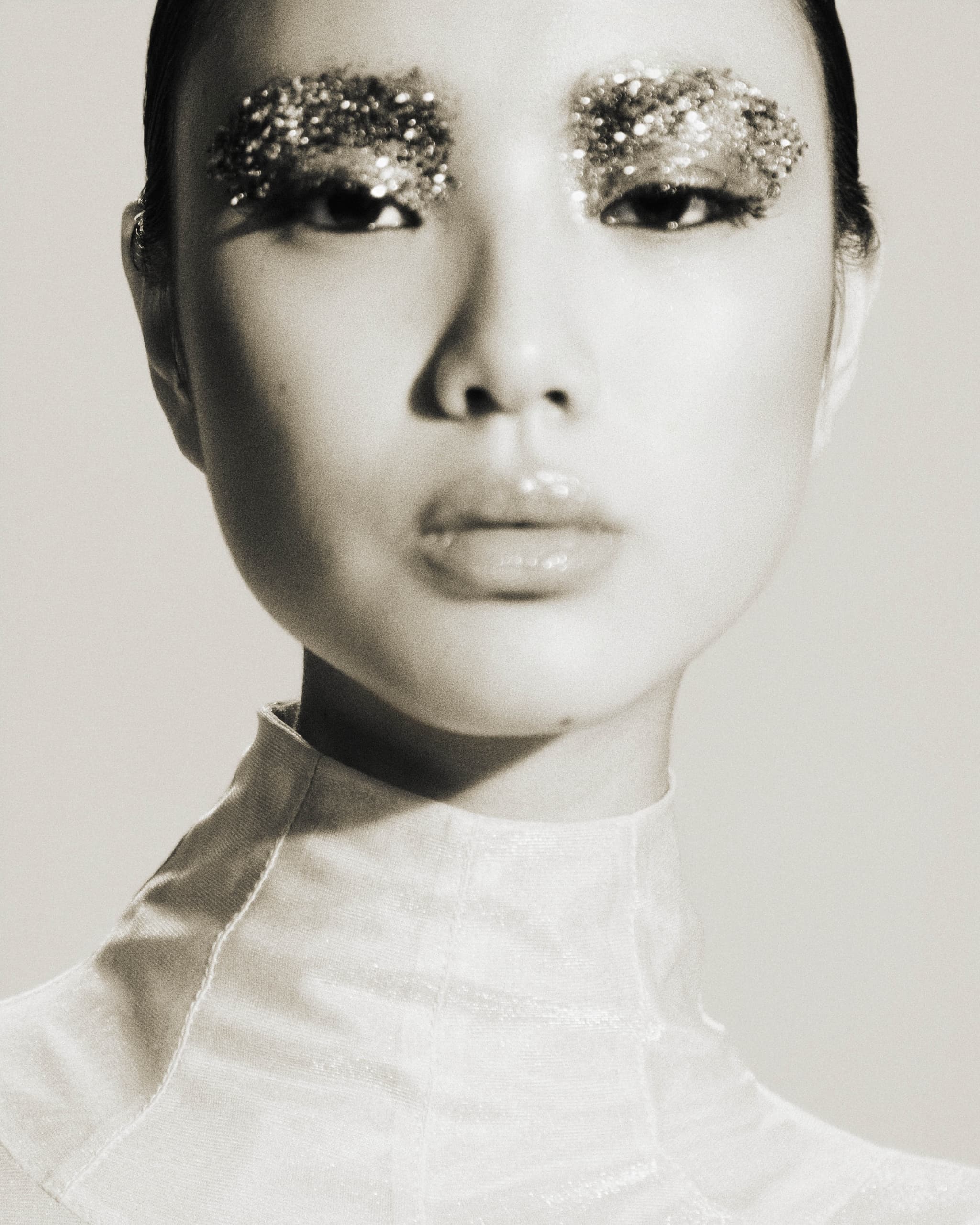 Model with glitter eyebrows in monochrome portrait, wearing high-neck top.