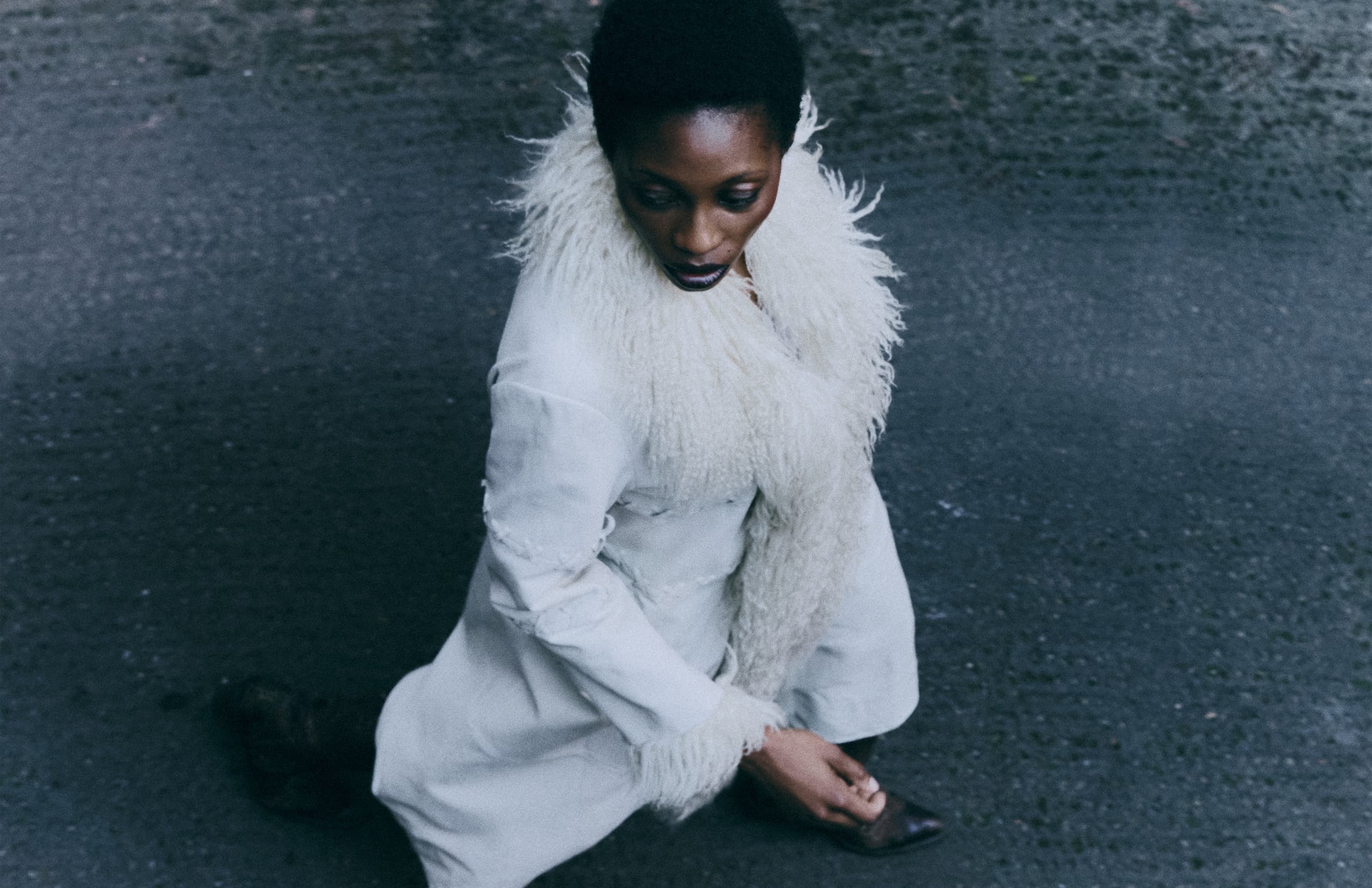 Woman in white coat and fur scarf kneels on textured pavement, looking down.