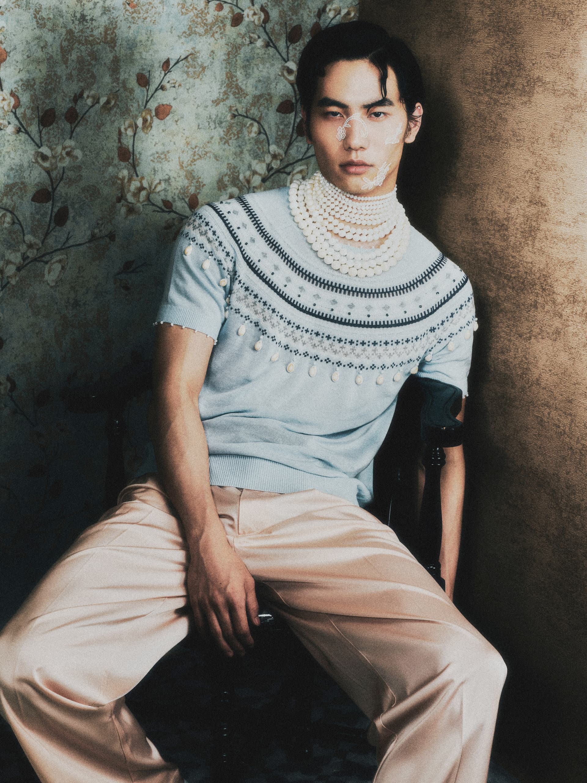 Stylish man wearing pearl-adorned sweater and beige pants in vintage setting.