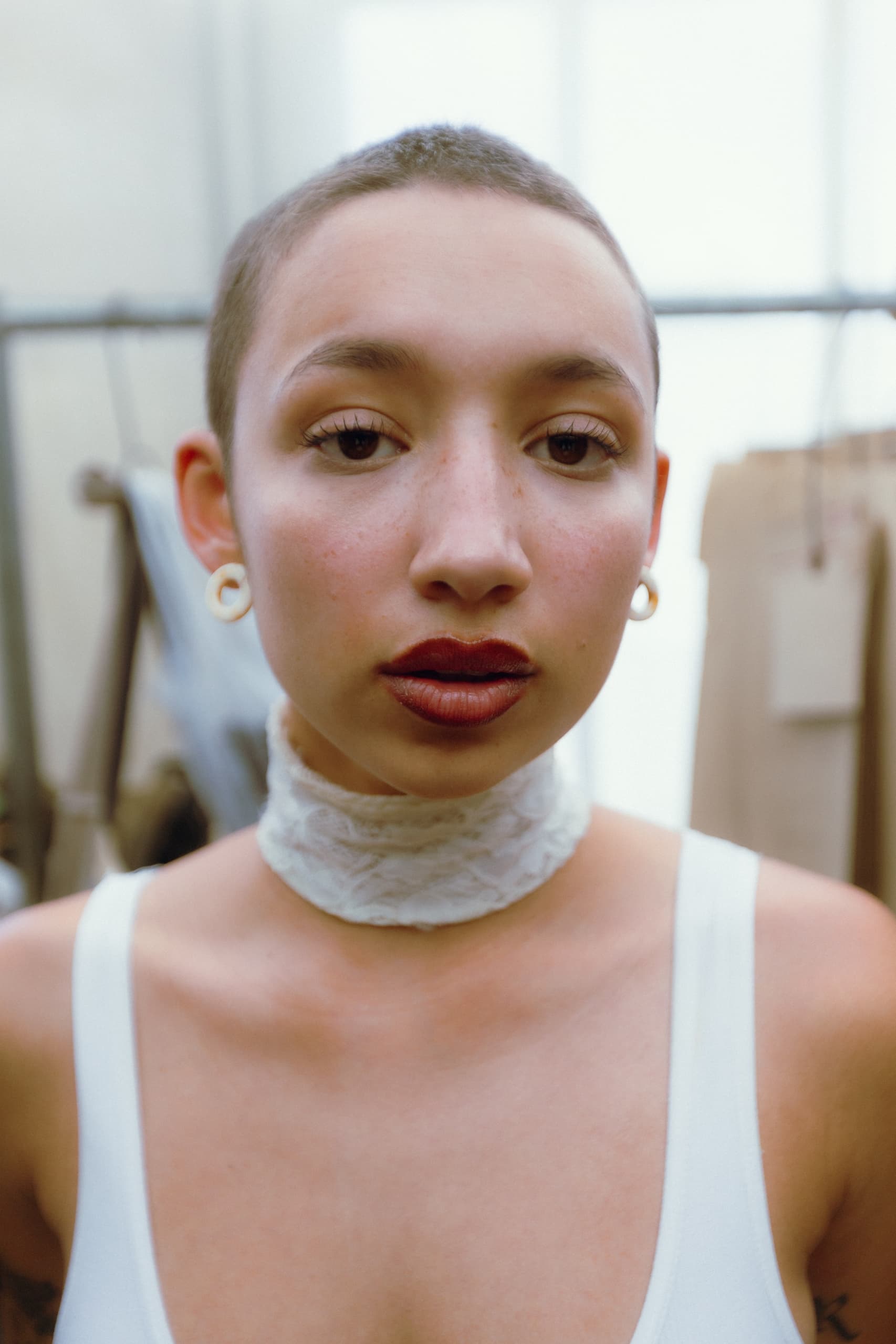 Person with shaved head, wearing lace choker and earrings, in a minimalistic setting.