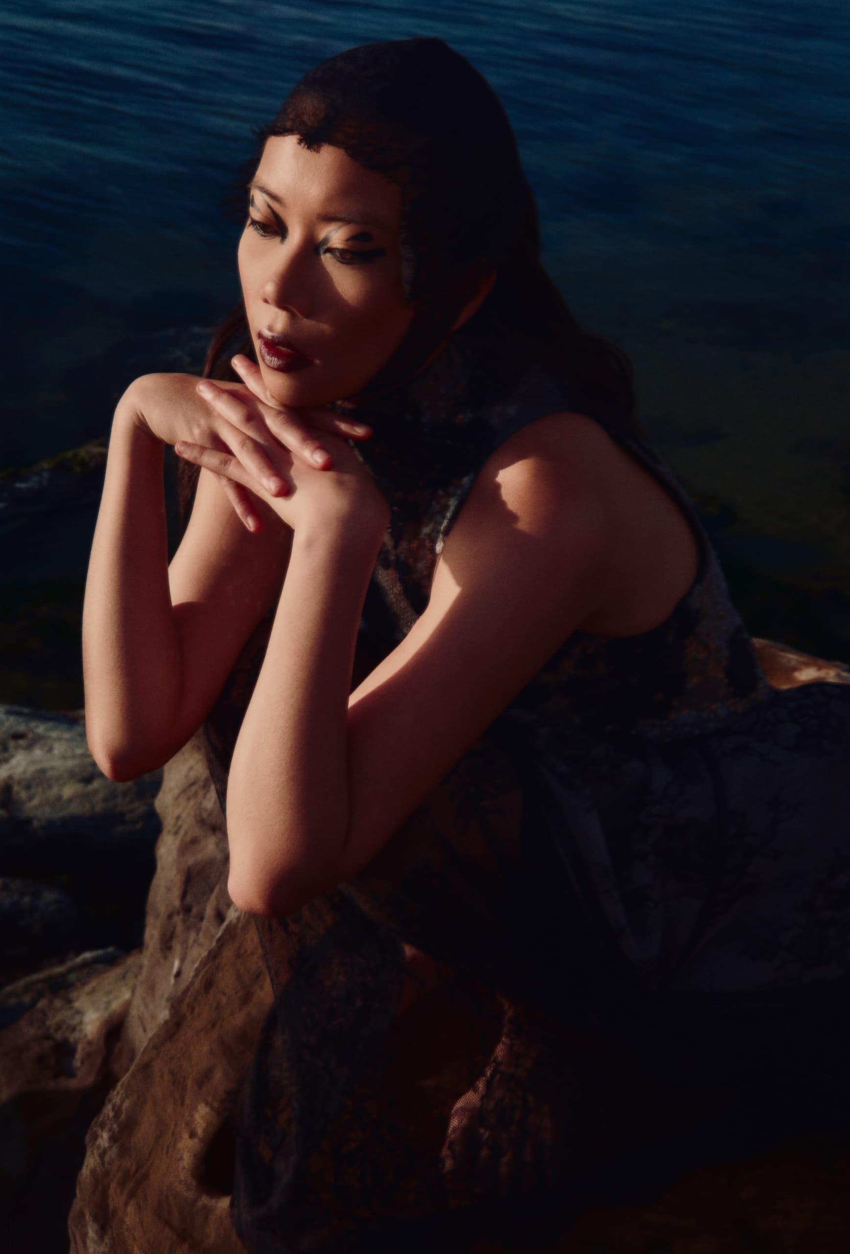 Person by the water in artistic pose, wearing dark lace attire, deep in thought.